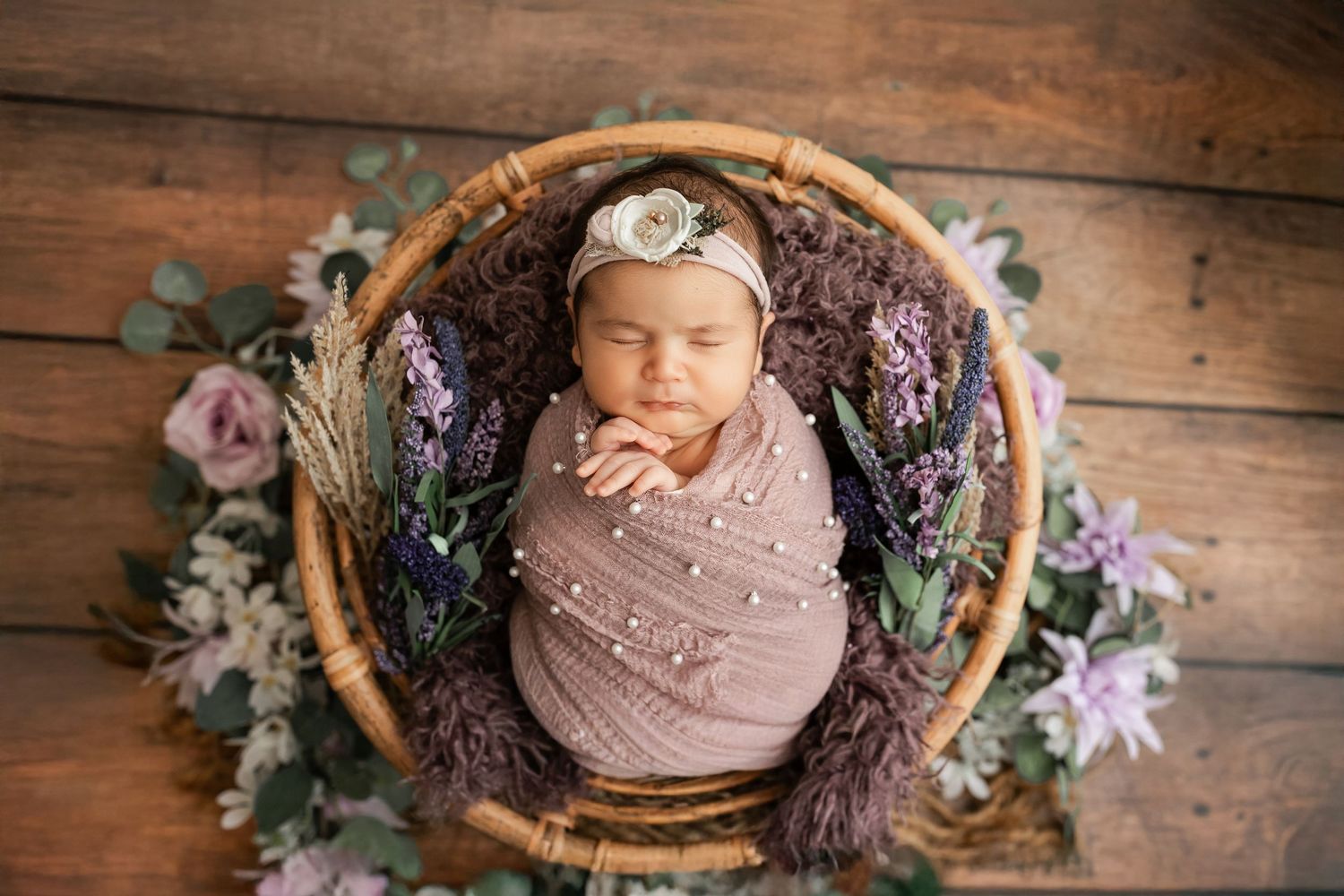 Newborn baby girl swaddled in pink wrap sleeping in a wicker basket surrounded by purple and pink flowers.