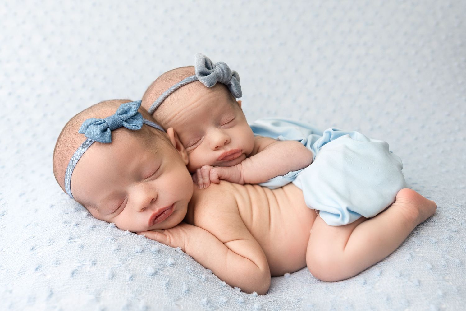 Sleeping newborn baby wearing a blue bow headband rests peacefully on white textured blanket in side profile pose.
