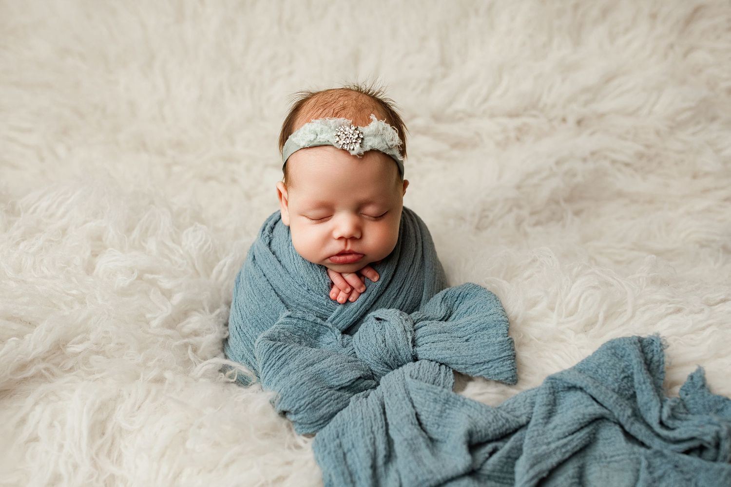 Sleeping newborn wrapped in soft blue swaddle blanket with lace headband on white furry backdrop.