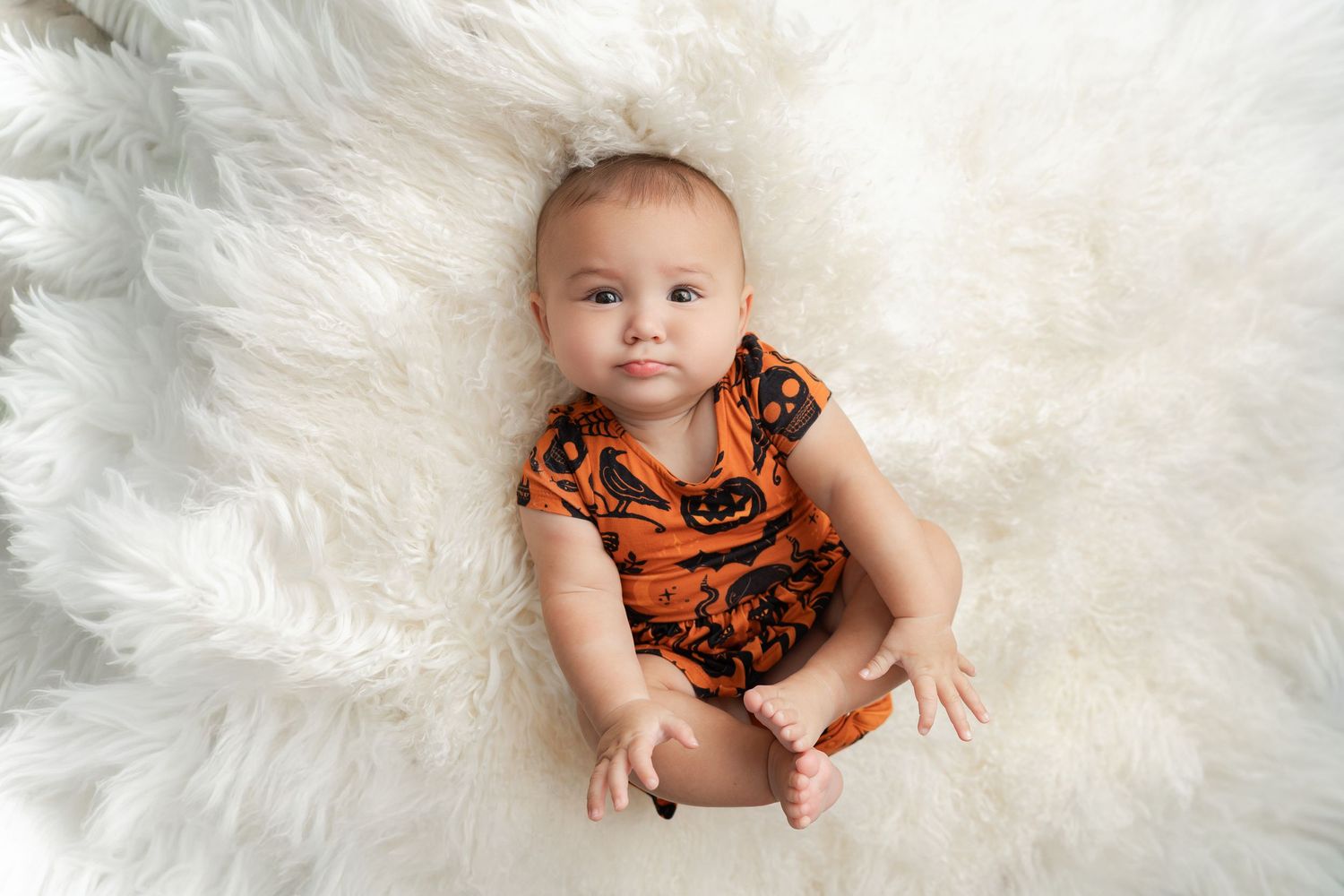 Baby in an orange and black patterned romper sits on a soft white fluffy sheepskin rug, looking up at the camera.