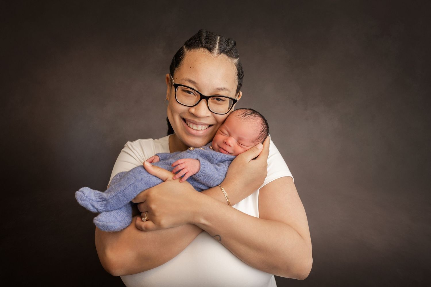 A smiling woman with braids and glasses holds a sleeping newborn wrapped in a blue swaddle against a dark background.
