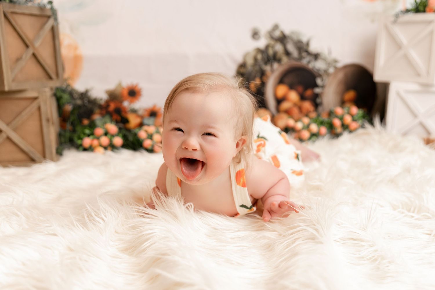 Happy baby laughing on a white fluffy rug surrounded by fall floral decorations in a bright studio setting.