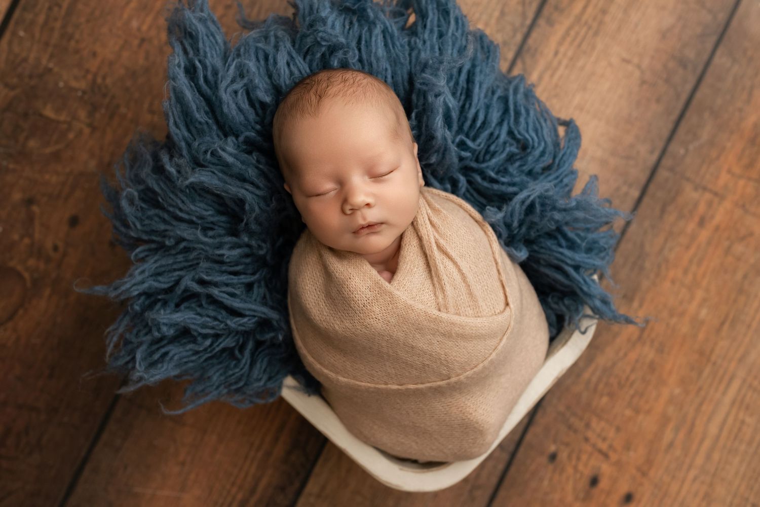 Peaceful newborn baby swaddled in tan wrap, sleeping on blue fluffy blanket in white bucket on wood floor.