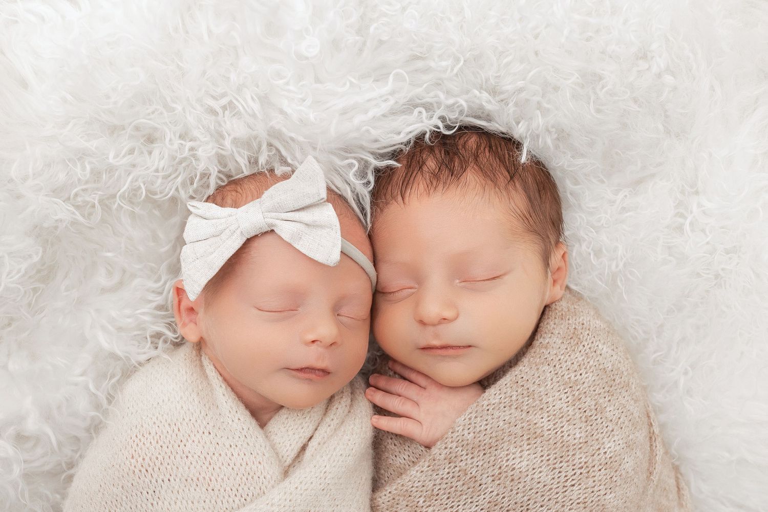 Two sleeping newborn babies wearing white bows rest together on soft white textured fabric.