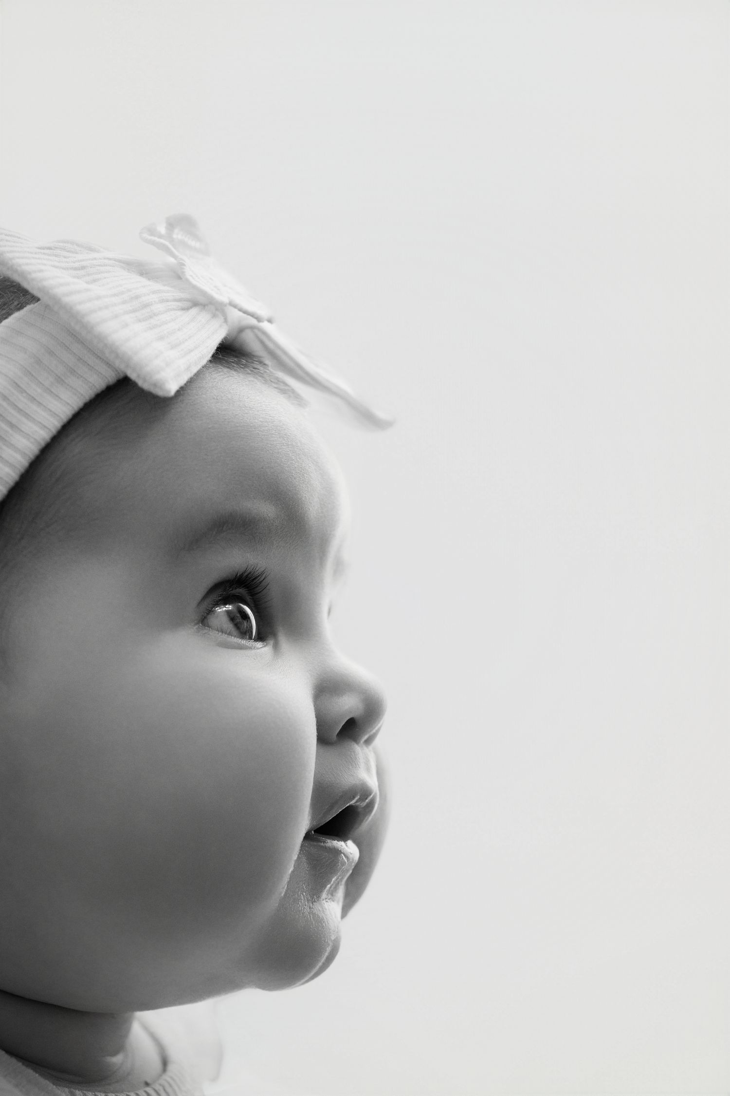 Black and white portrait of a cute baby girl wearing a bow headband, looking up with wide curious eyes.