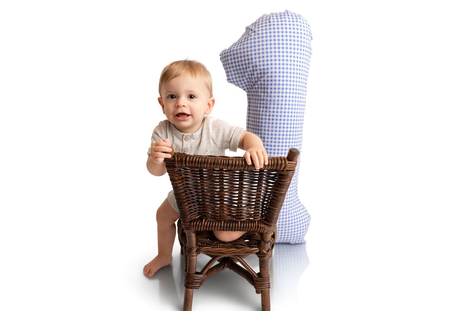 Baby standing behind a small wicker chair next to a blue gingham number one pillow, celebrating a first birthday against a white background.