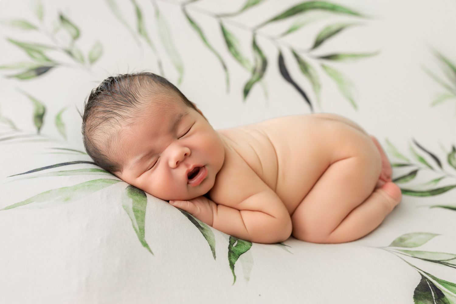 Newborn baby sleeping peacefully on a white floral blanket with green leaves in a professional photography studio.