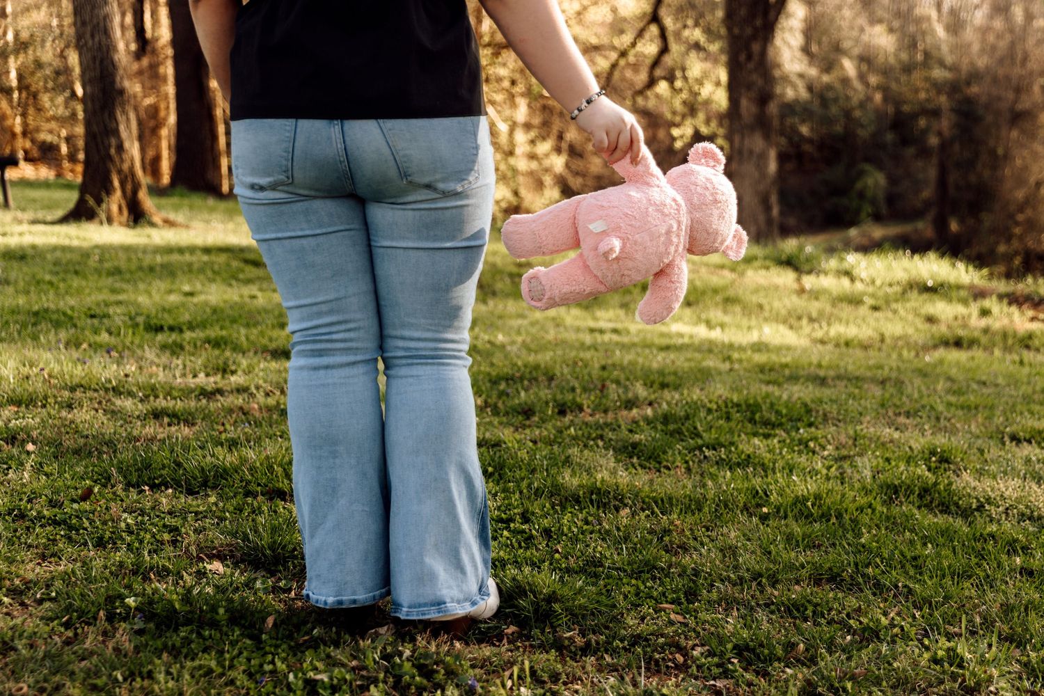 Child holding pink teddy bear standing on green grass in outdoor park setting.
