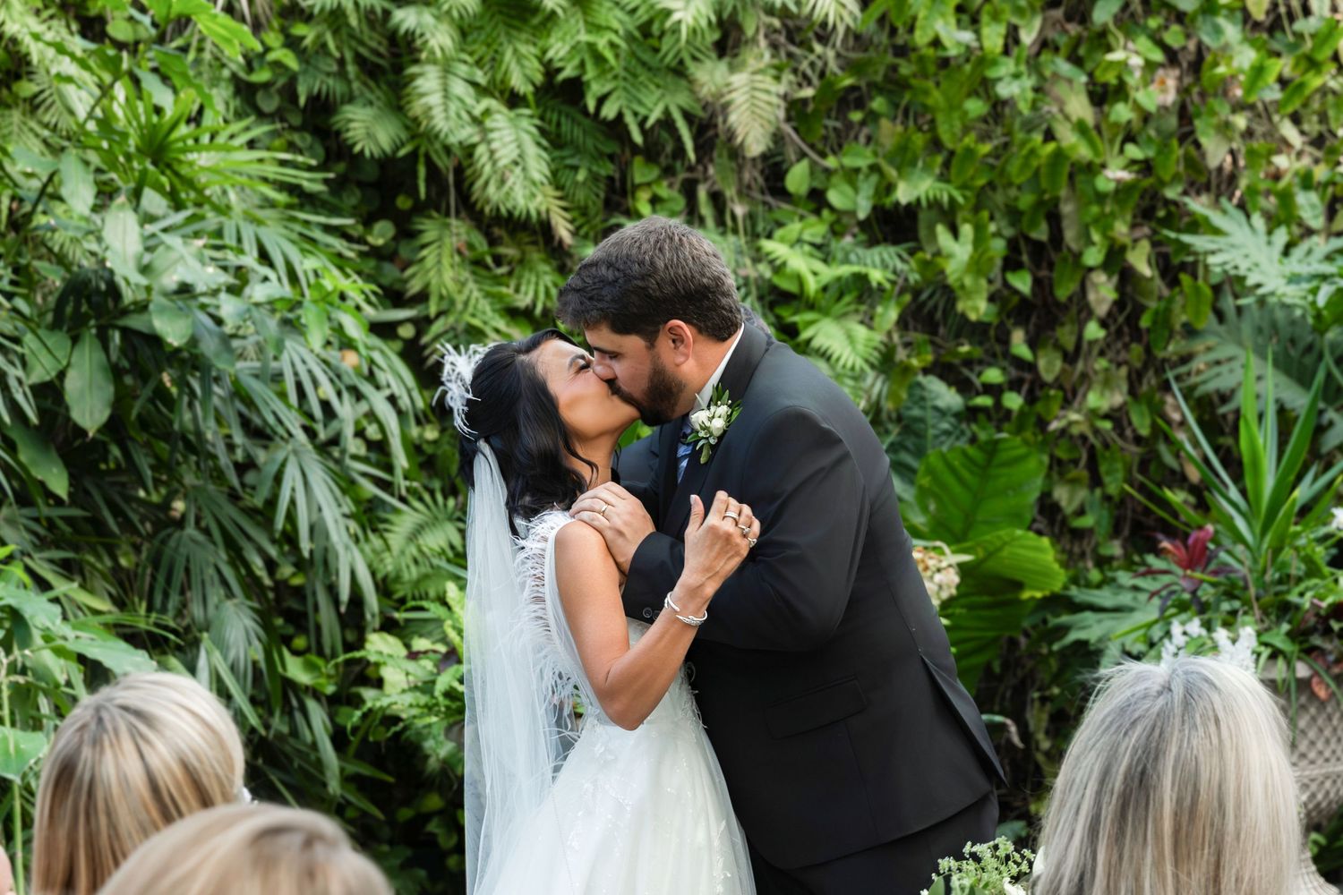 Bride and groom share first kiss at lush tropical greenery wedding ceremony altar surrounded by guests.