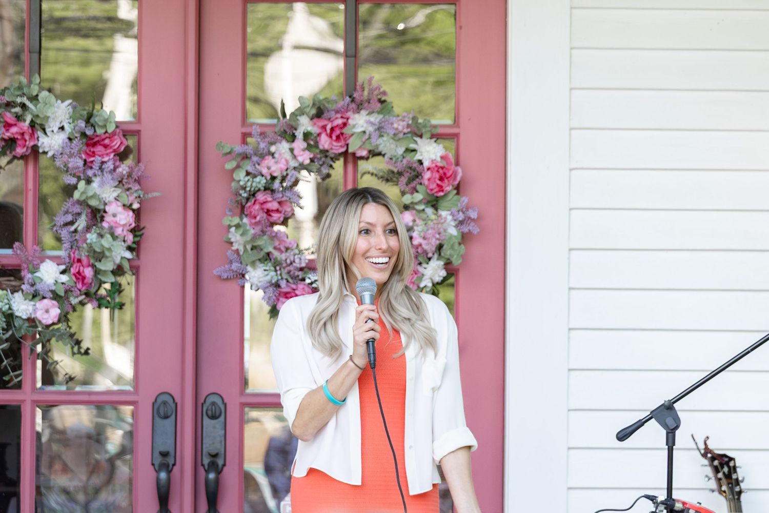 Woman with blonde hair speaks into microphone outdoors near pink door with floral wreath, wearing orange and white outfit.