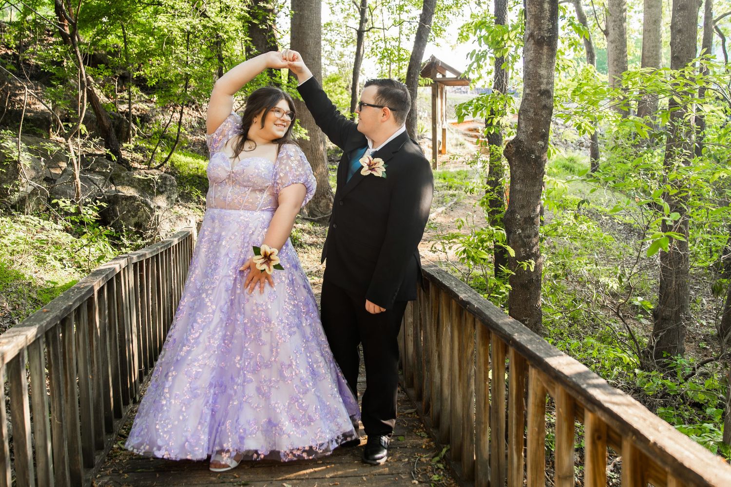 Couple in formal wear dancing on a wooden bridge surrounded by lush green trees during prom season.
