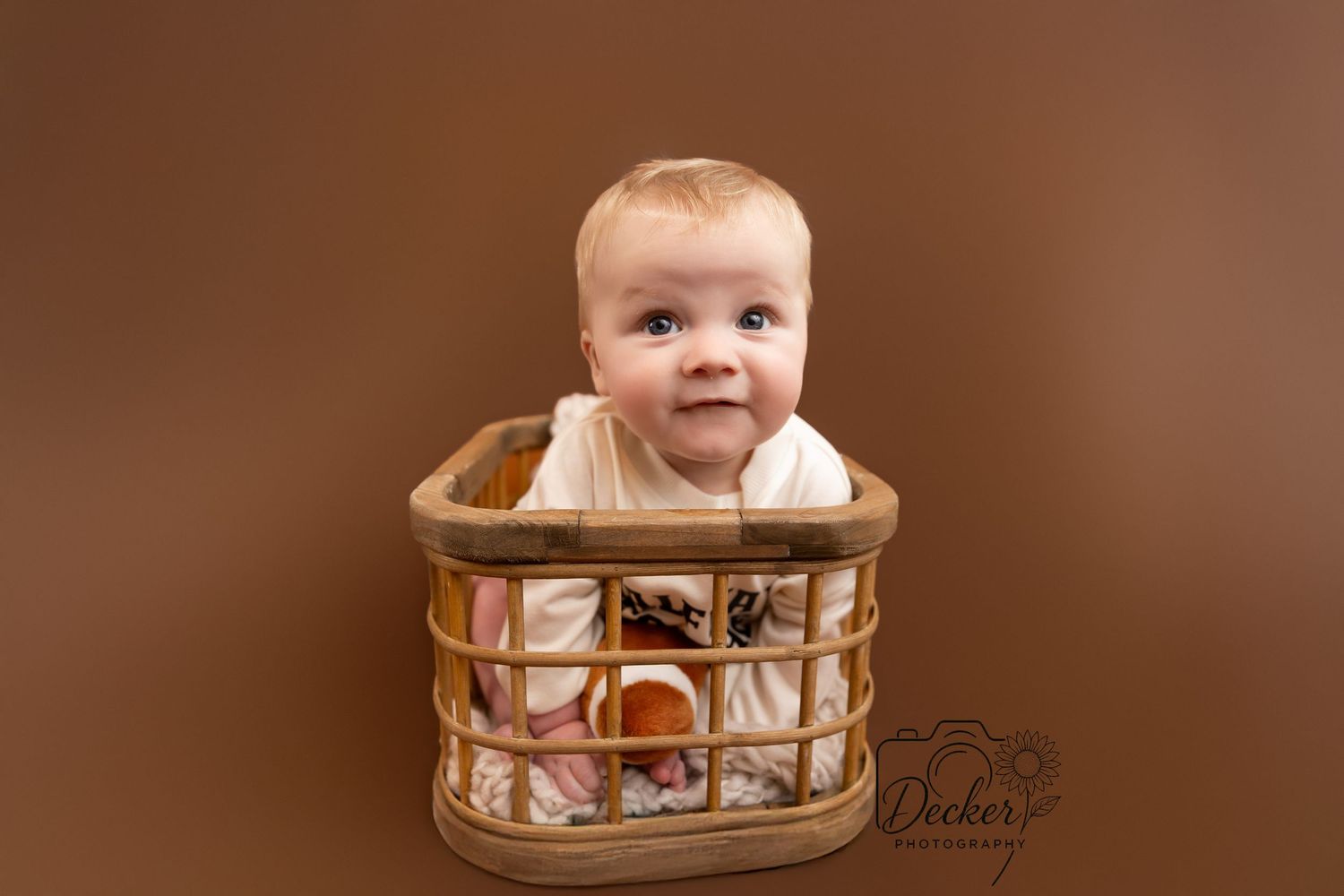 A baby smiles while sitting in a wooden high chair against a brown backdrop.