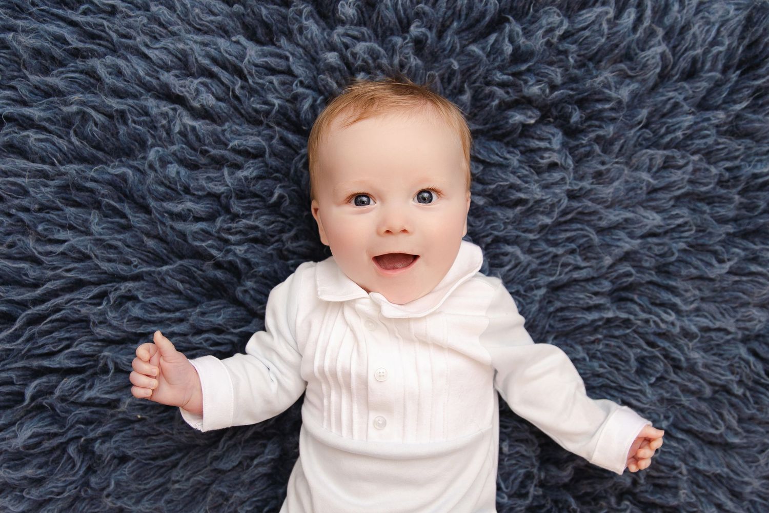 A baby wearing a white sweater lies on a dark blue shag carpet in a series of candid poses.