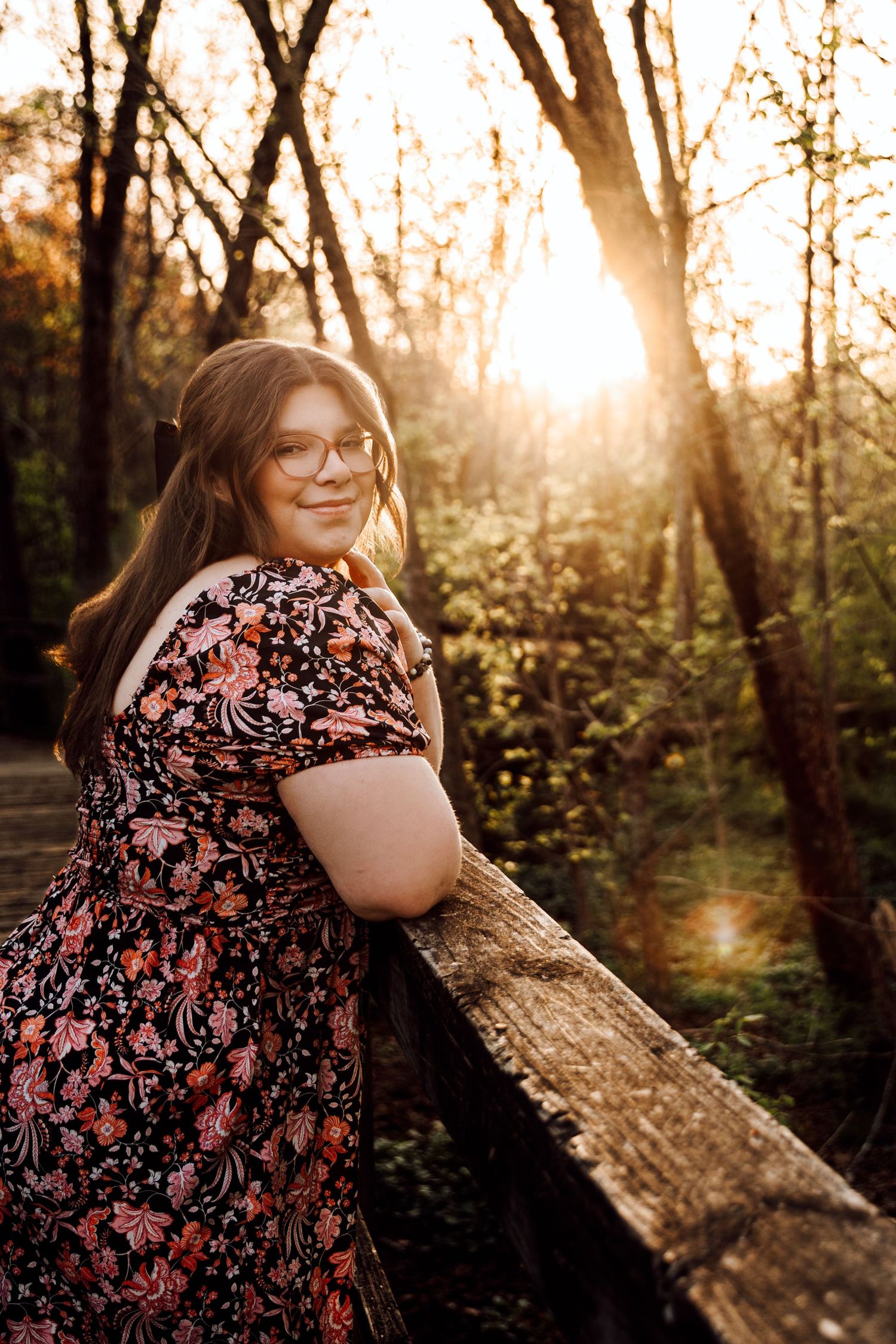 Young woman in floral dress leaning on wooden railing at golden hour sunset in forest.