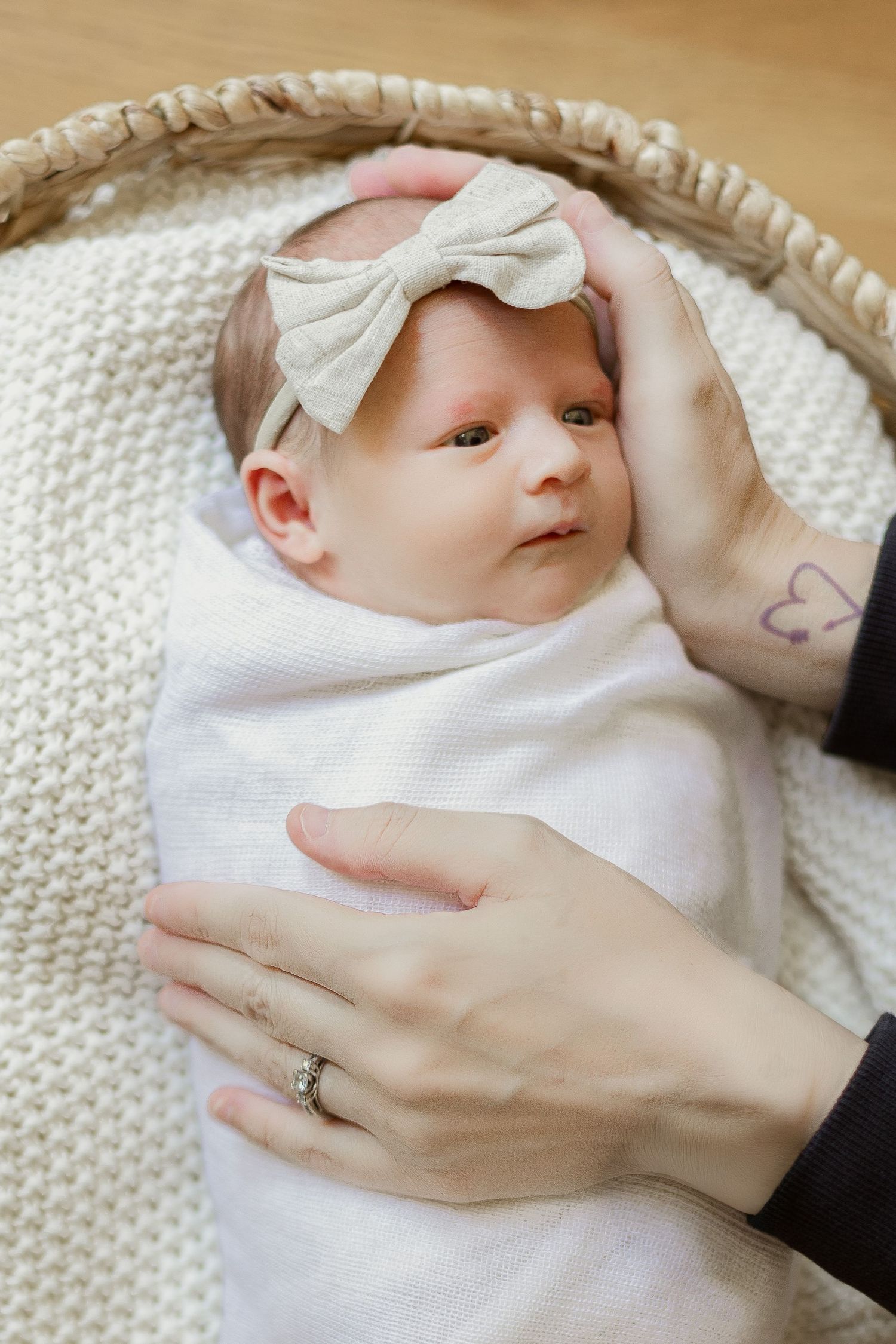 A baby wearing a white bow headband and swaddle lies on a knitted blanket while gentle hands reach to comfort.