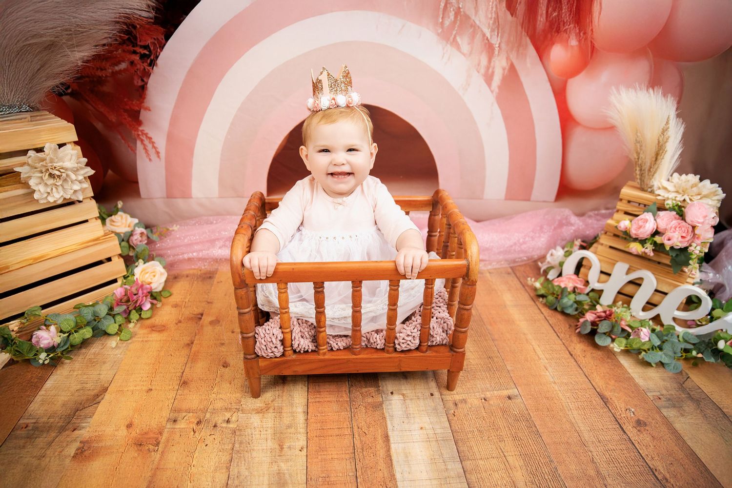 Pink rainbow backdrop with wooden crib and floral decorations on hardwood floor in studio setting.