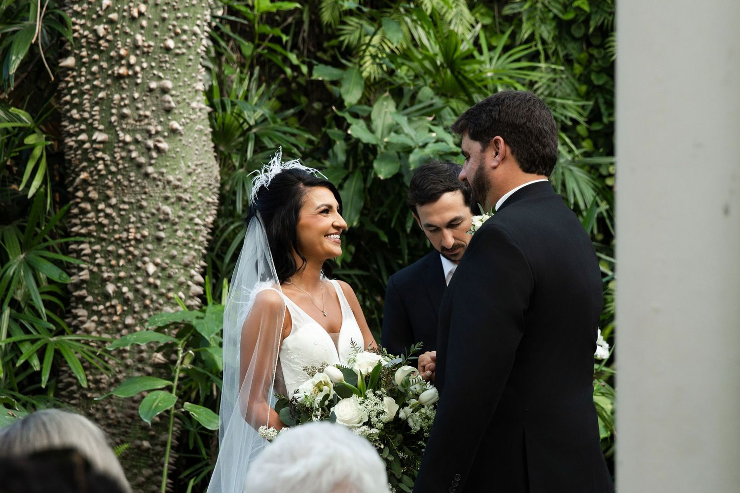 Bride in white gown and veil holds bouquet while exchanging vows with groom in black suit at outdoor wedding ceremony.