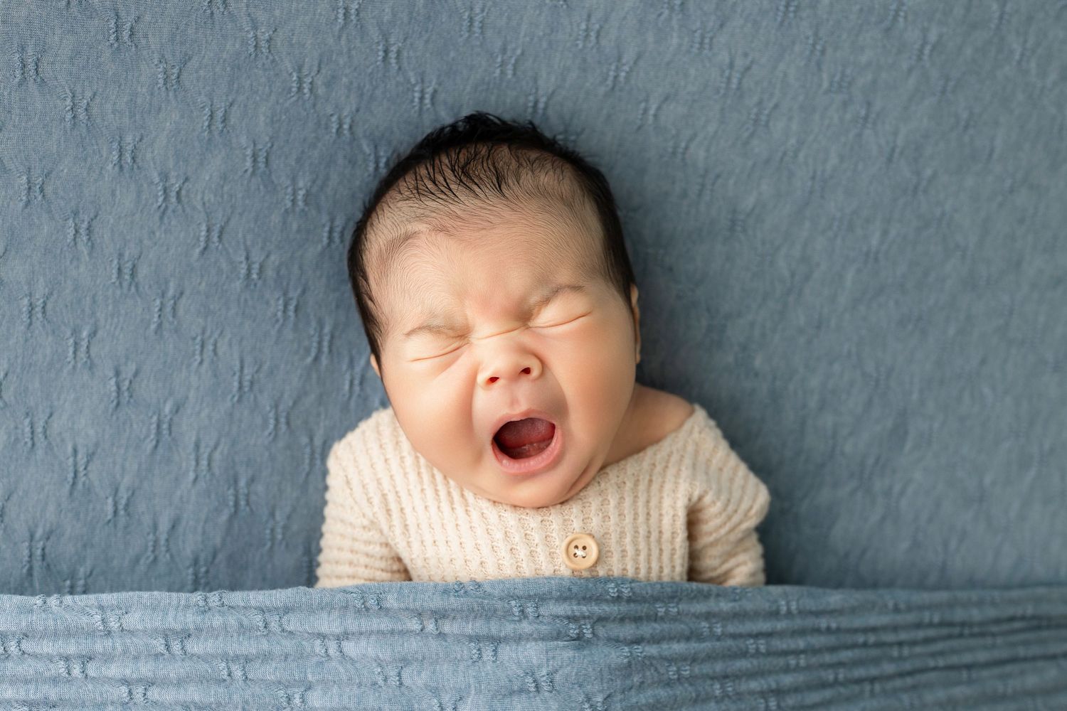 Adorable newborn baby yawning in a cream knit outfit, lying on a soft blue textured blanket.
