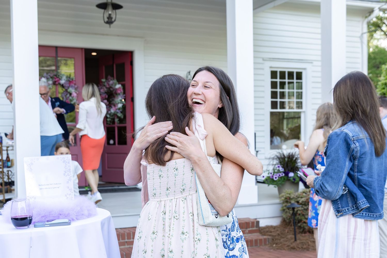 Two women share a joyful hug outdoors at a garden party near a white house with a red door.