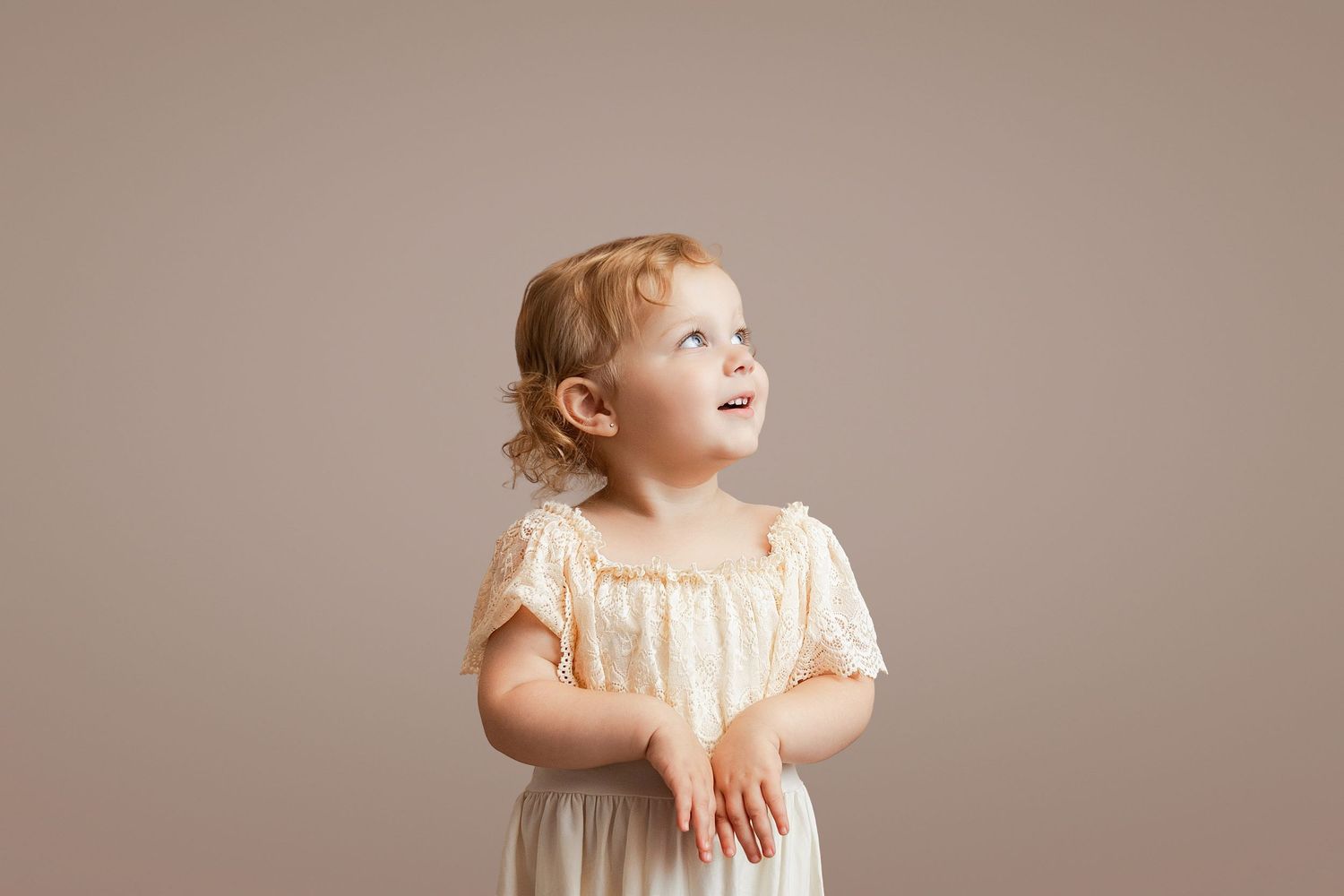 Adorable toddler girl in cream dress looking up curiously against neutral beige background.