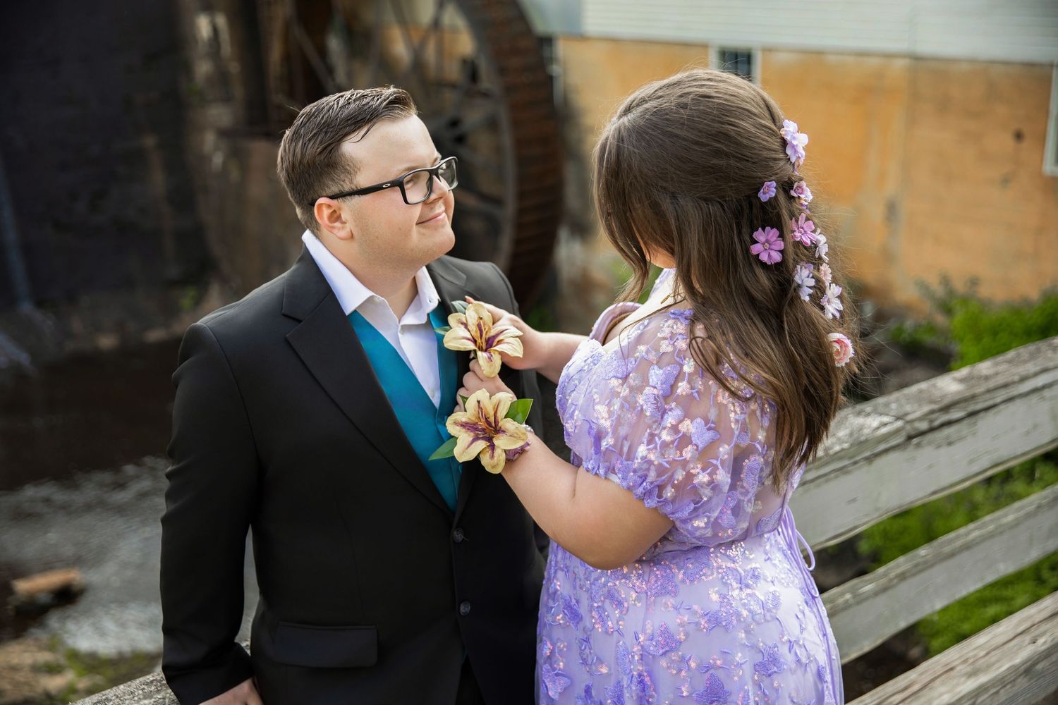 Couple in formal wear pinning a boutonniere, woman in lavender dress with floral hair accessories.