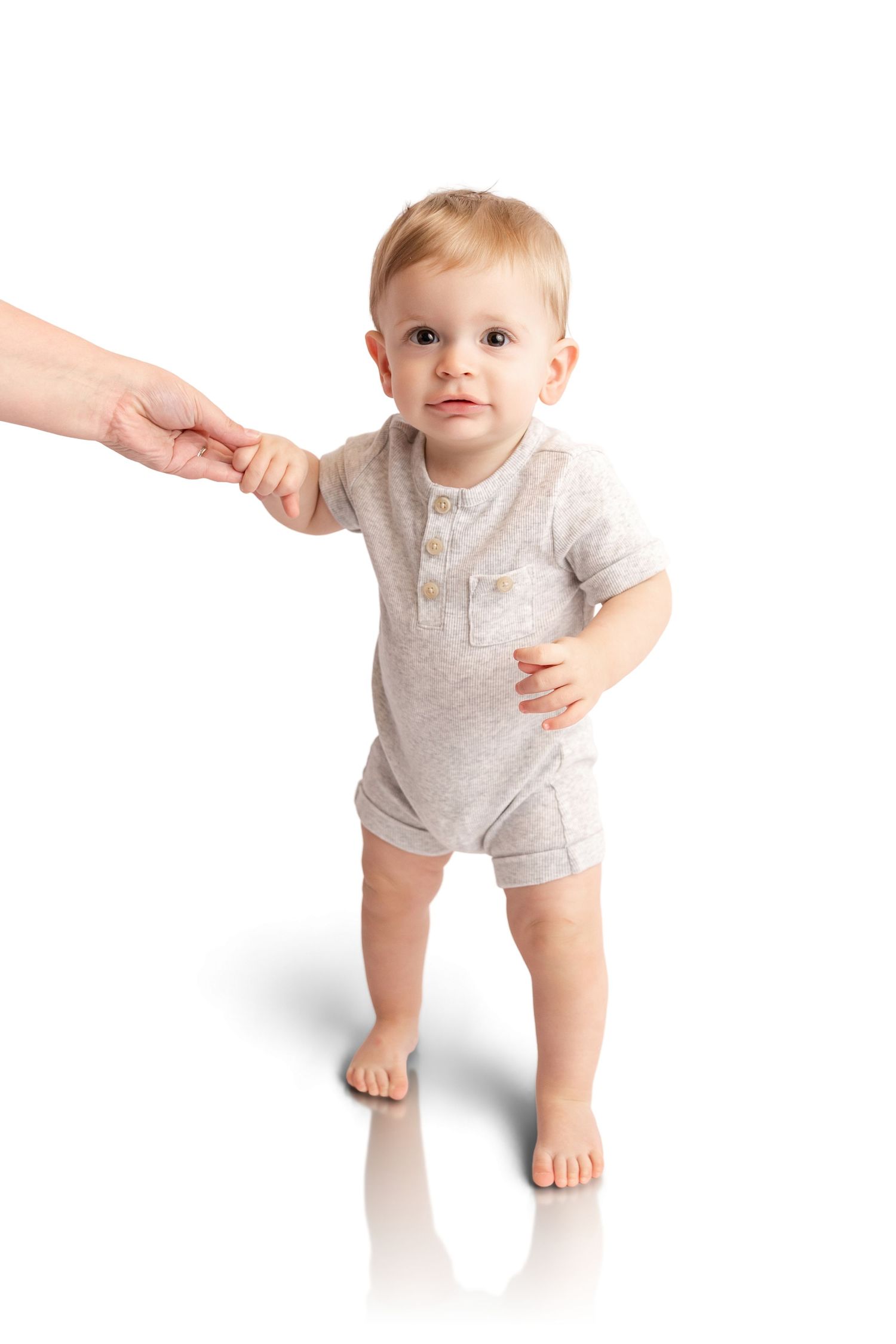 Baby in a gray short-sleeve romper stands barefoot on a white background while holding an adult's hand during first steps.