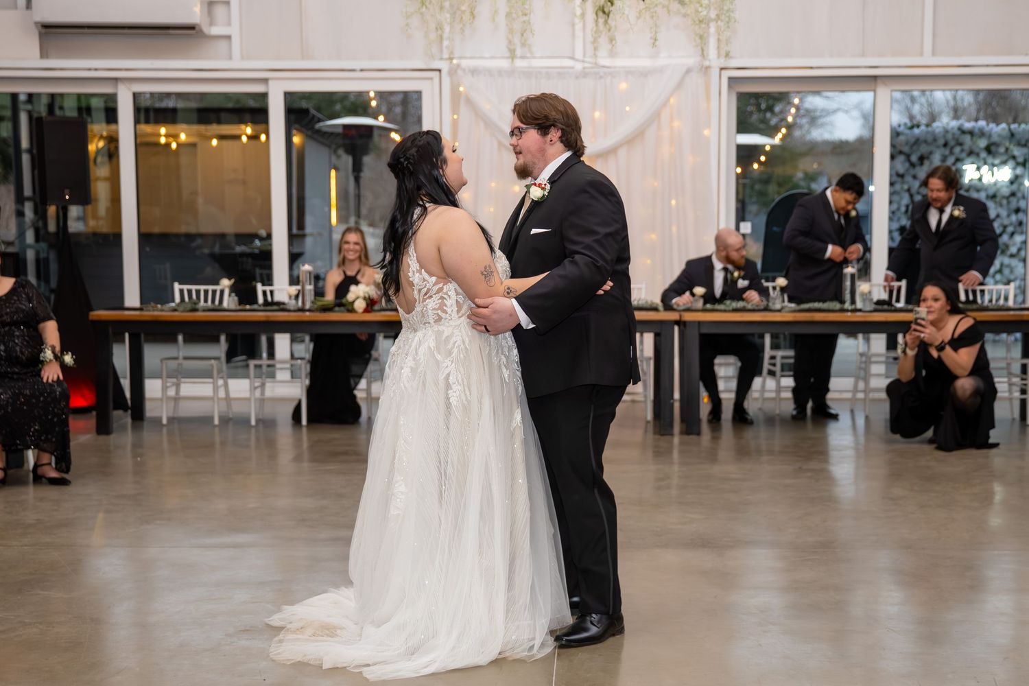 Newlyweds share first dance in elegant reception venue with guests seated at tables behind them.