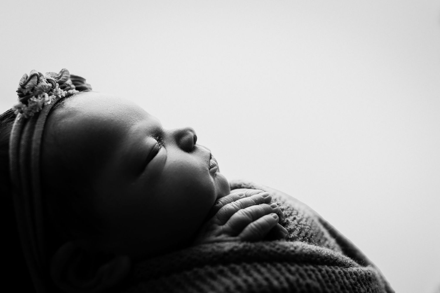 Black and white profile portrait of a sleeping newborn baby wearing a floral headband, resting peacefully on a soft knit blanket.