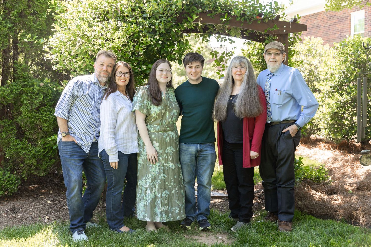 Six people smiling together outdoors in a lush garden setting for a family portrait photo.