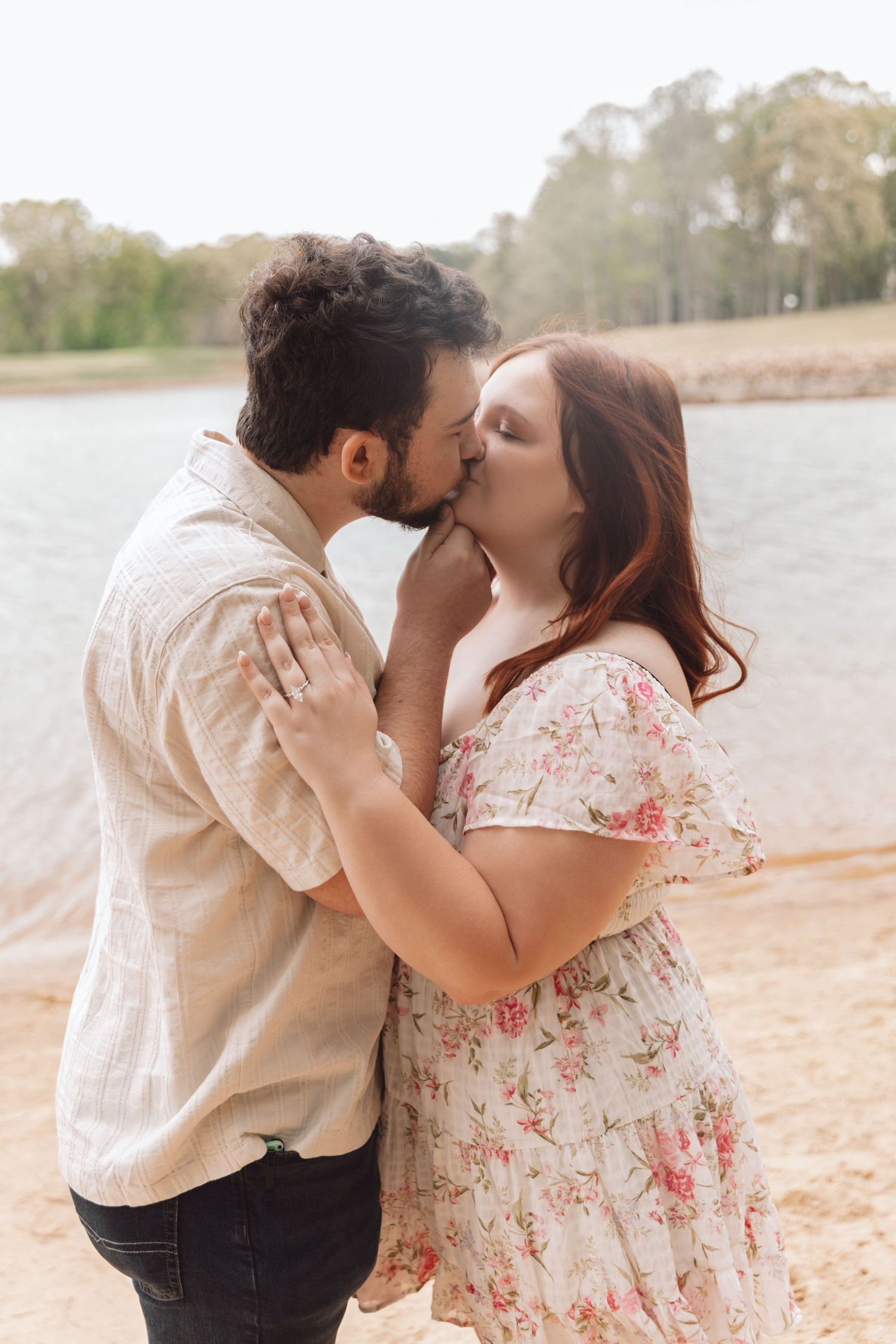 Couple sharing a romantic kiss by a lake, woman in floral dress and man in cream shirt embracing outdoors.