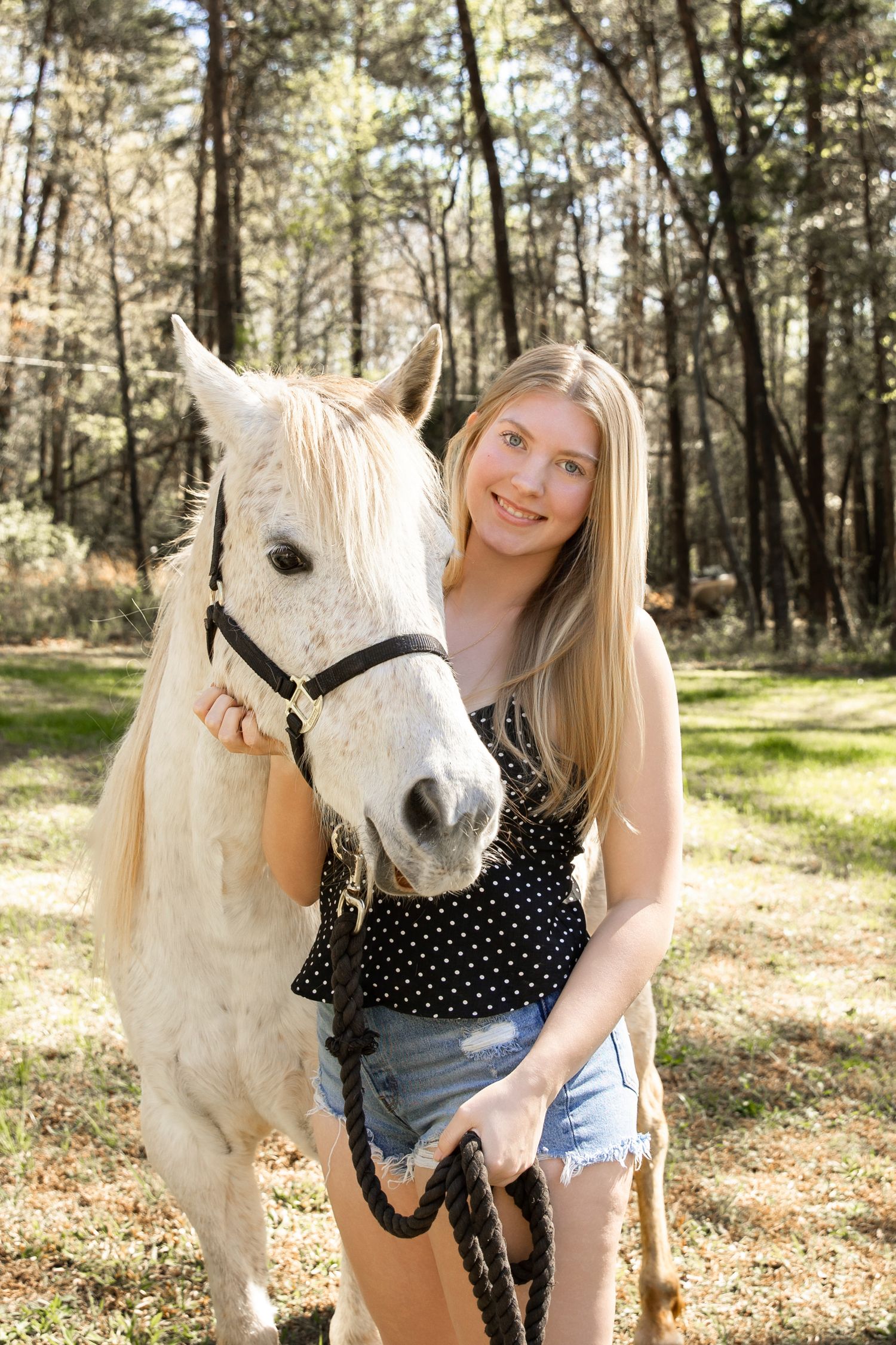 Young blonde woman smiling beside a white horse with a black halter in a sunny forest clearing.