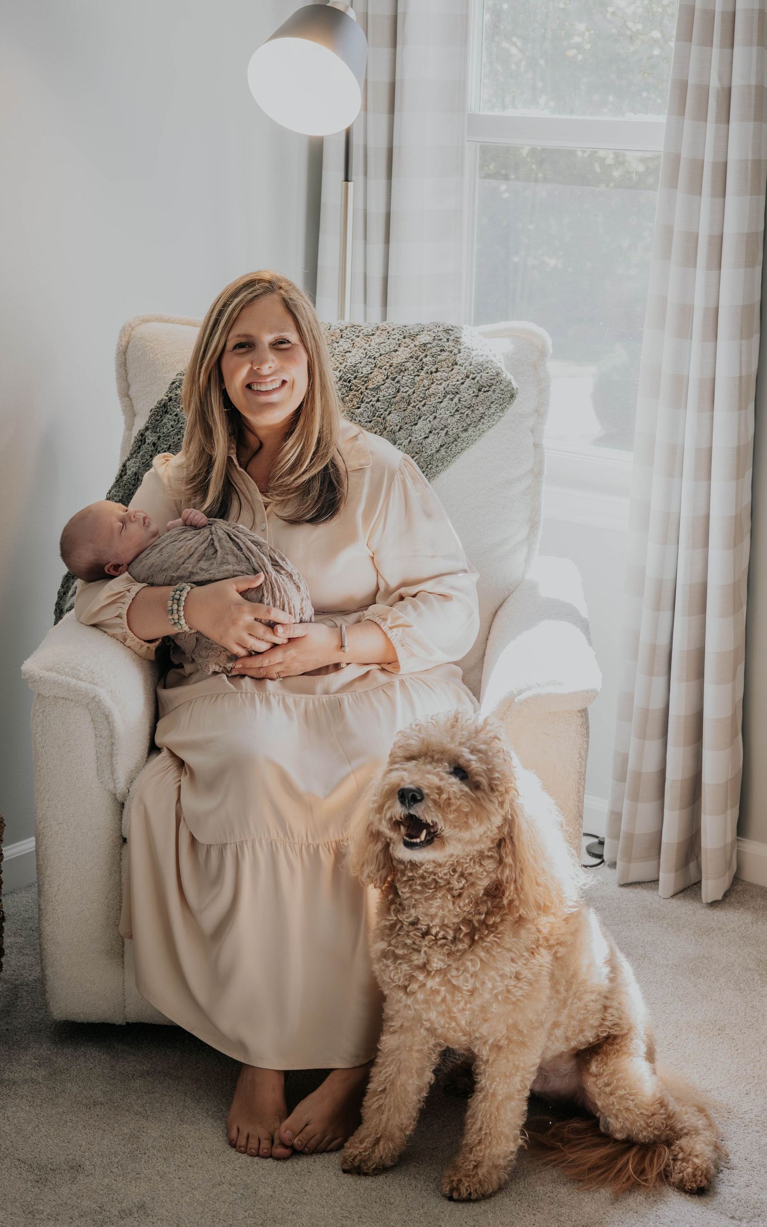 A cozy indoor scene with a fluffy goldendoodle dog sitting beside someone in a cream-colored dress on a plush chair.