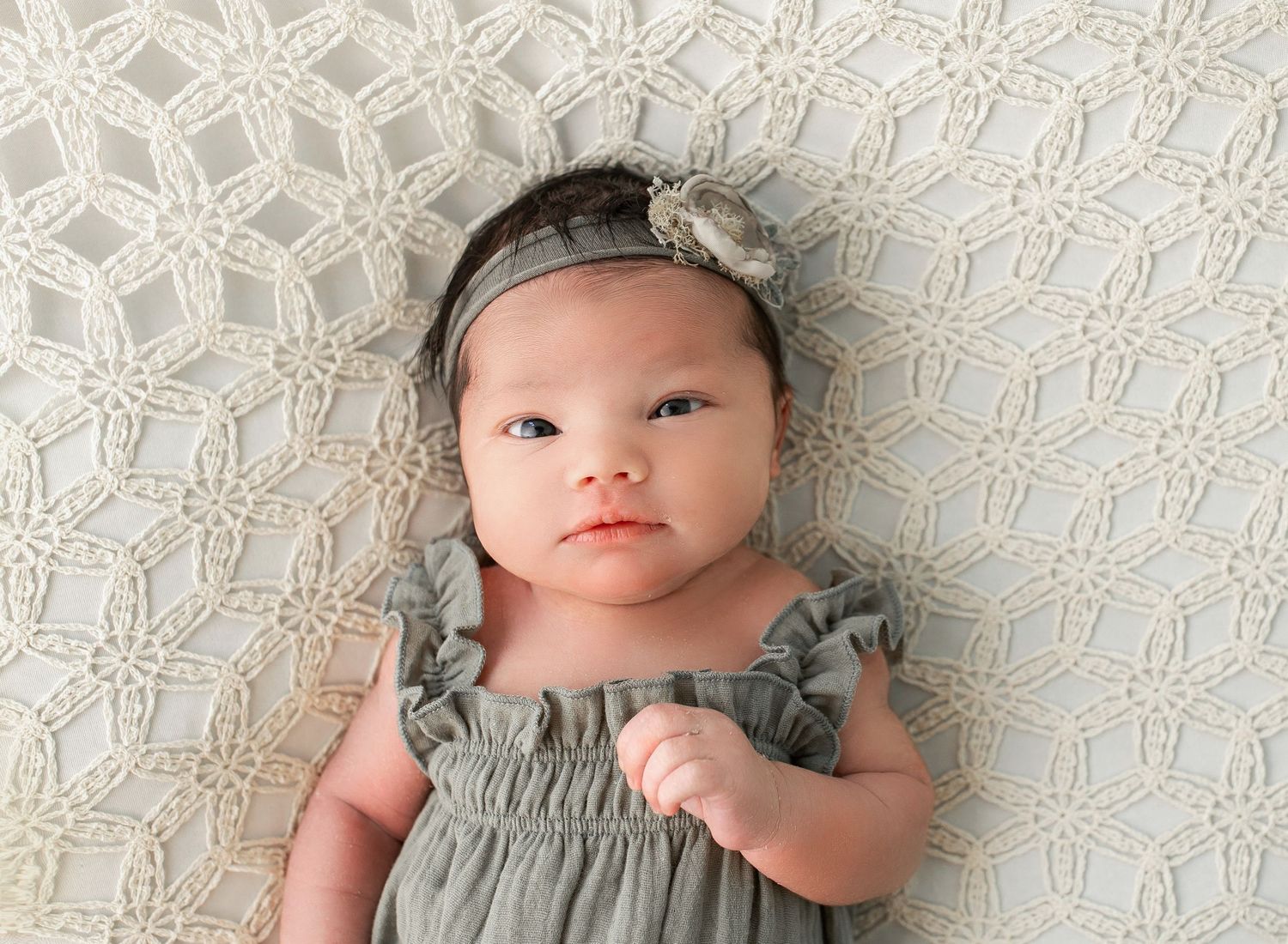 Newborn baby girl in sage green romper and floral headband lying on white lace blanket.