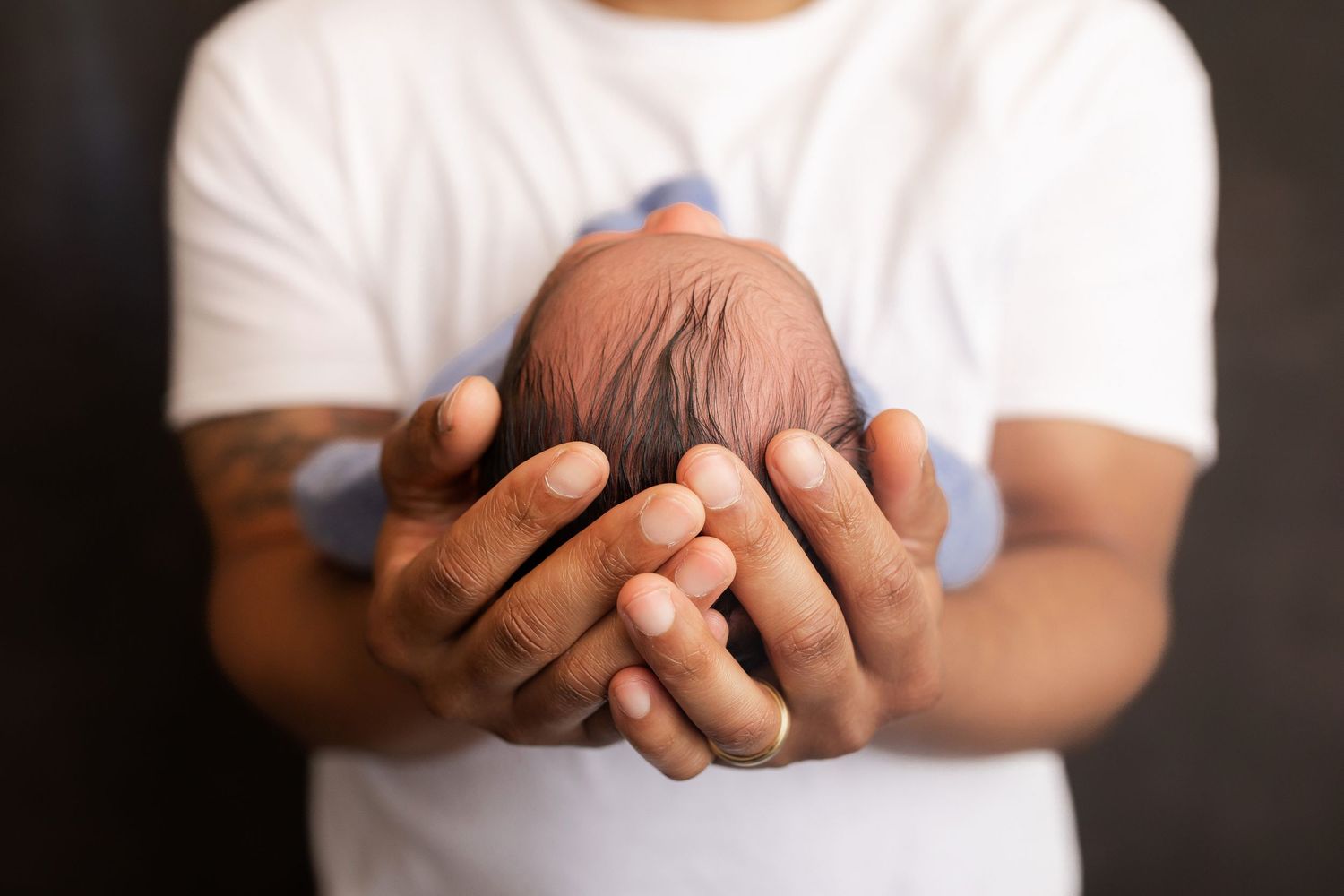 A father in a white t-shirt gently cradles his newborn baby in both hands against a dark background.