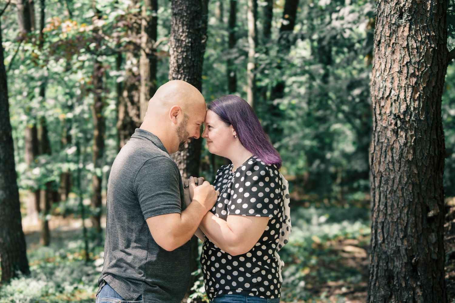 A couple shares an intimate moment together while standing in a sunlit wooded forest area.