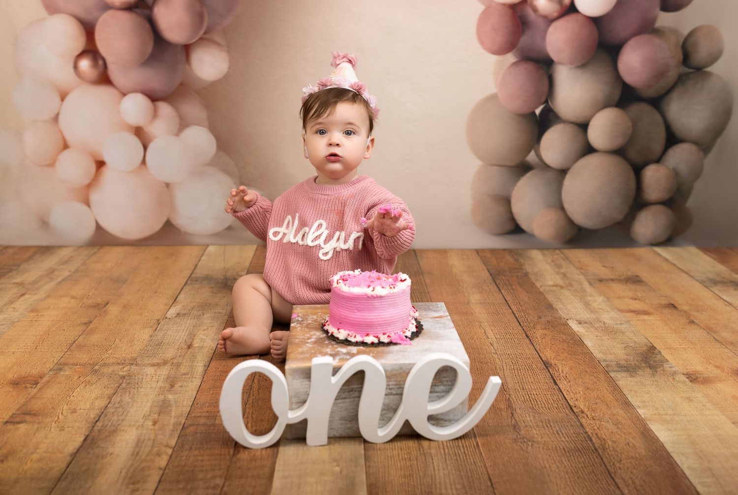 First birthday celebration setup with pink cake, large white 'one' sign, and blush balloon arch on wooden floor.