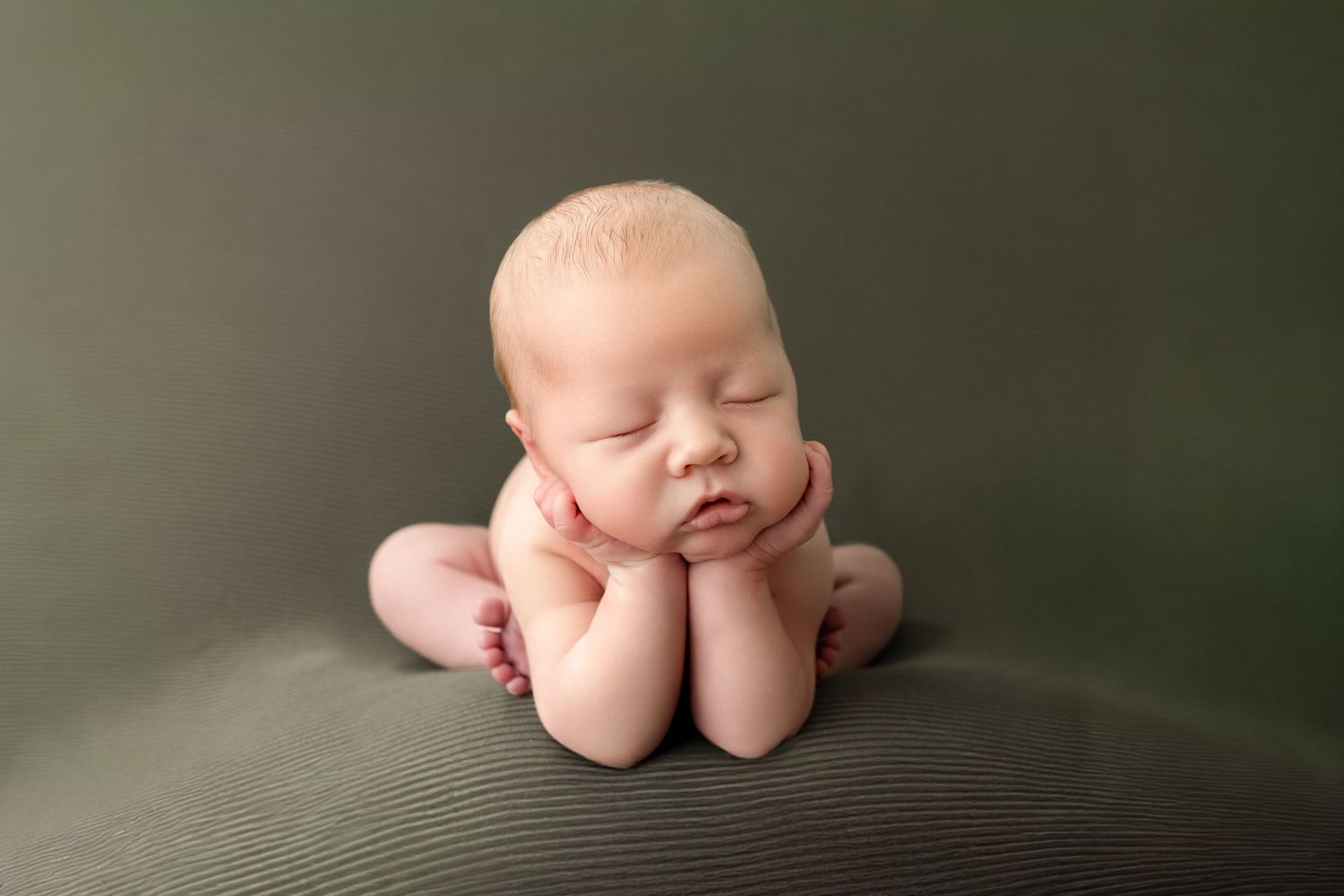 Newborn baby sleeping peacefully with hands under chin in a classic froggy pose on a green backdrop.