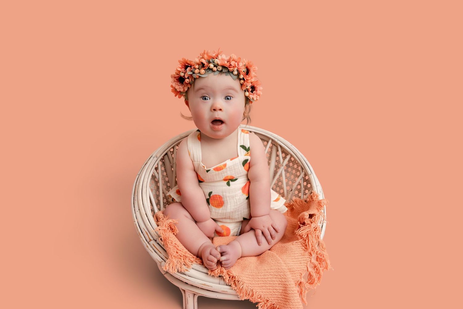 A cute baby girl in an orange floral romper and flower crown sits in a wicker chair on a peach background.