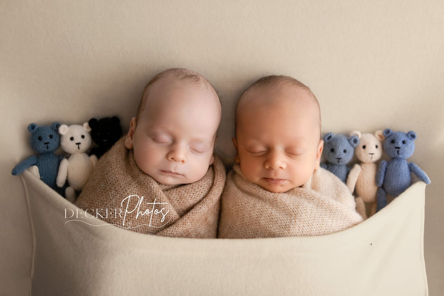 Two sleeping newborn twins wrapped in beige swaddles rest in a bowl surrounded by small knitted teddy bears in a studio photo.