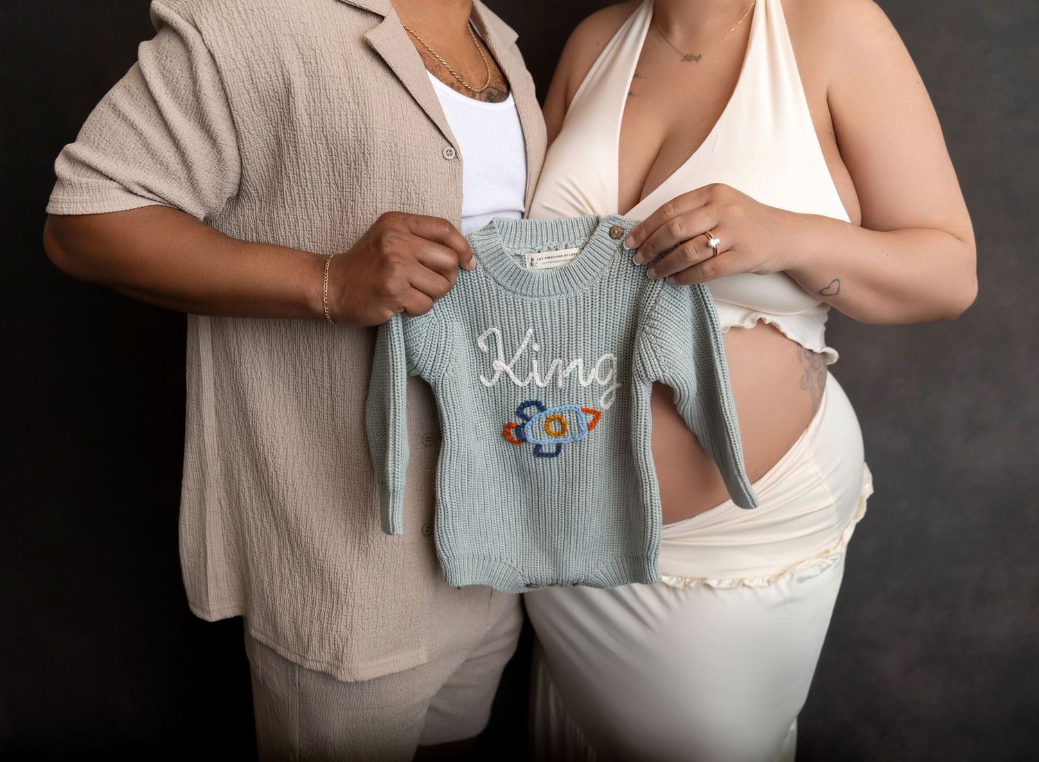 Couple holding a light blue knit baby onesie embroidered with 