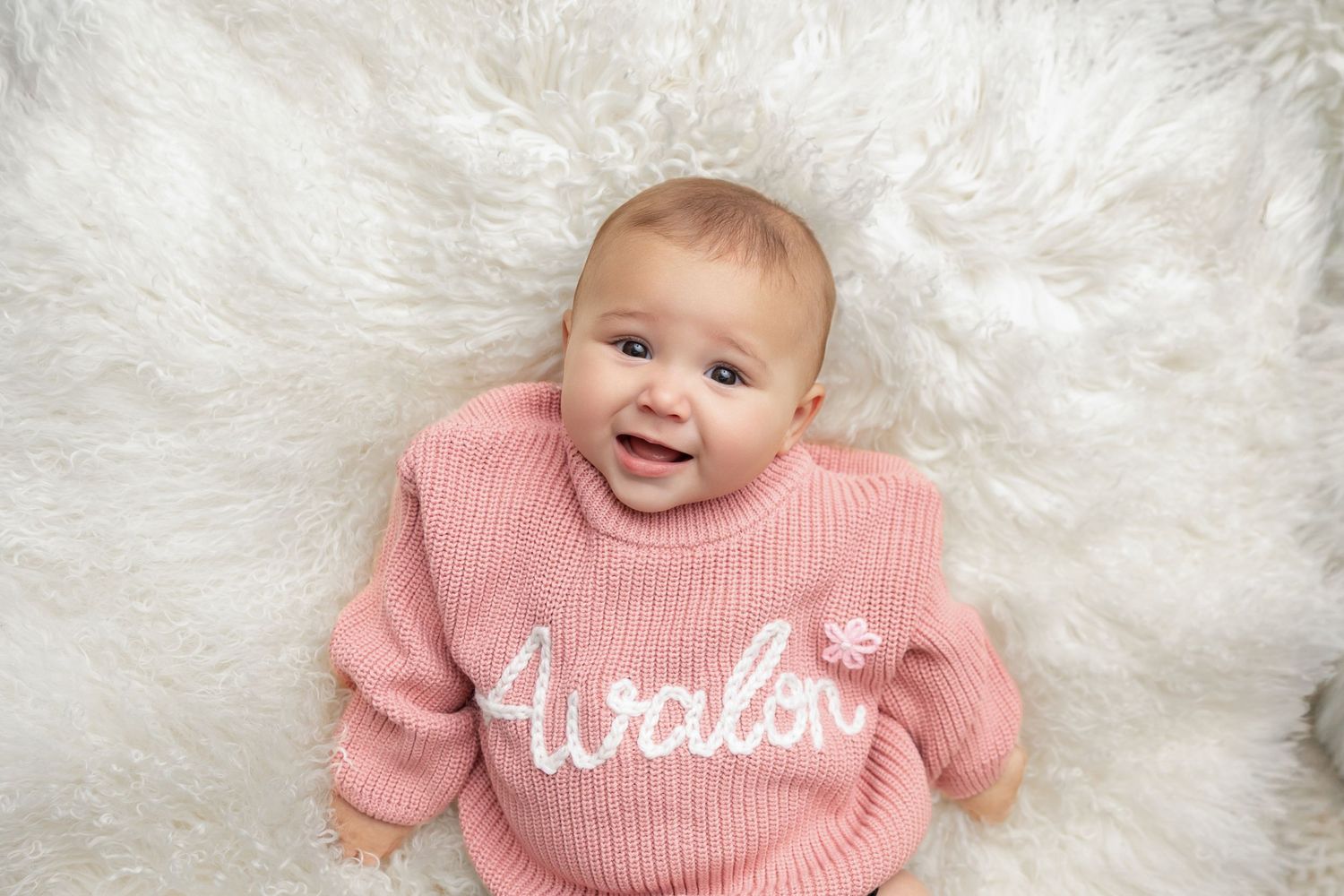 Smiling baby in a pink knit sweater with the name Avalon embroidered in white, lying on a soft white fluffy sheepskin rug.