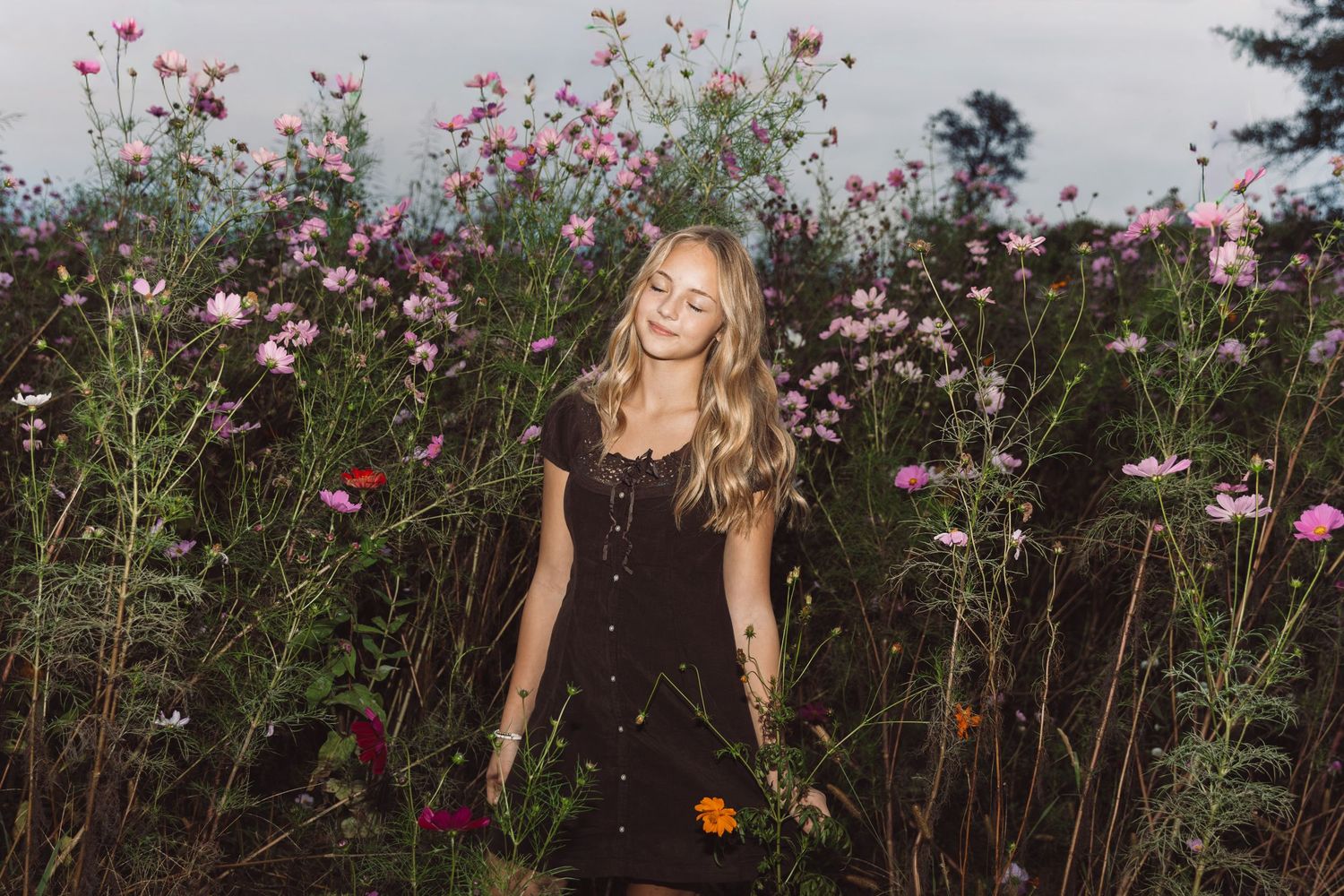 Smiling young woman with long wavy blonde hair in a black button dress standing among pink cosmos wildflowers in a meadow at golden hour.