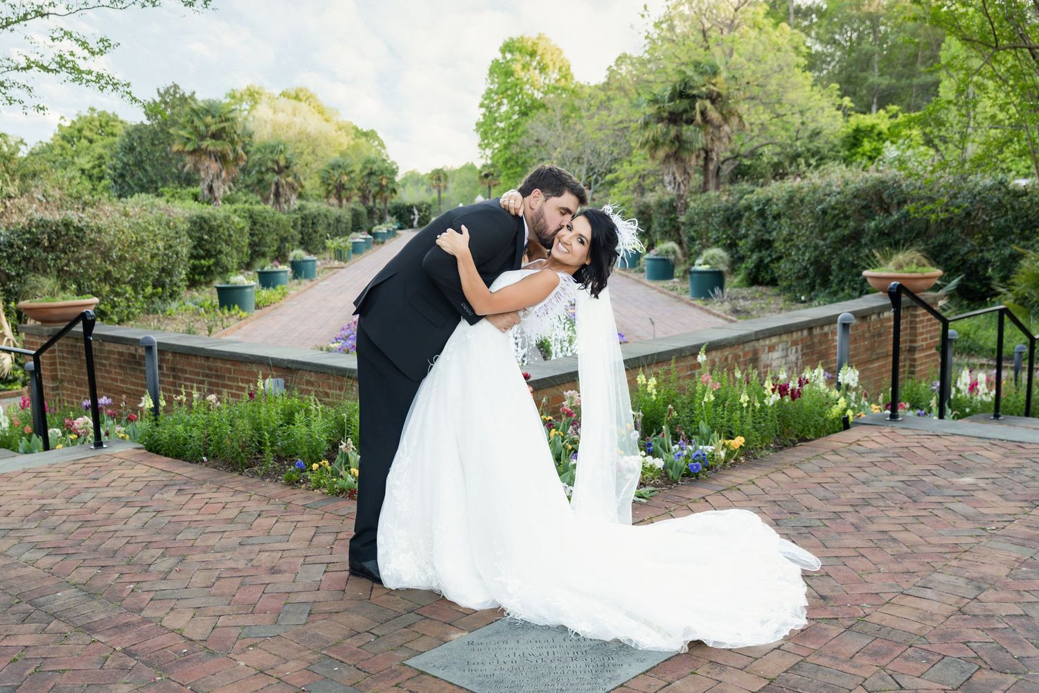 Bride and groom share a romantic dip kiss in a lush garden setting, surrounded by colorful flowers and greenery.