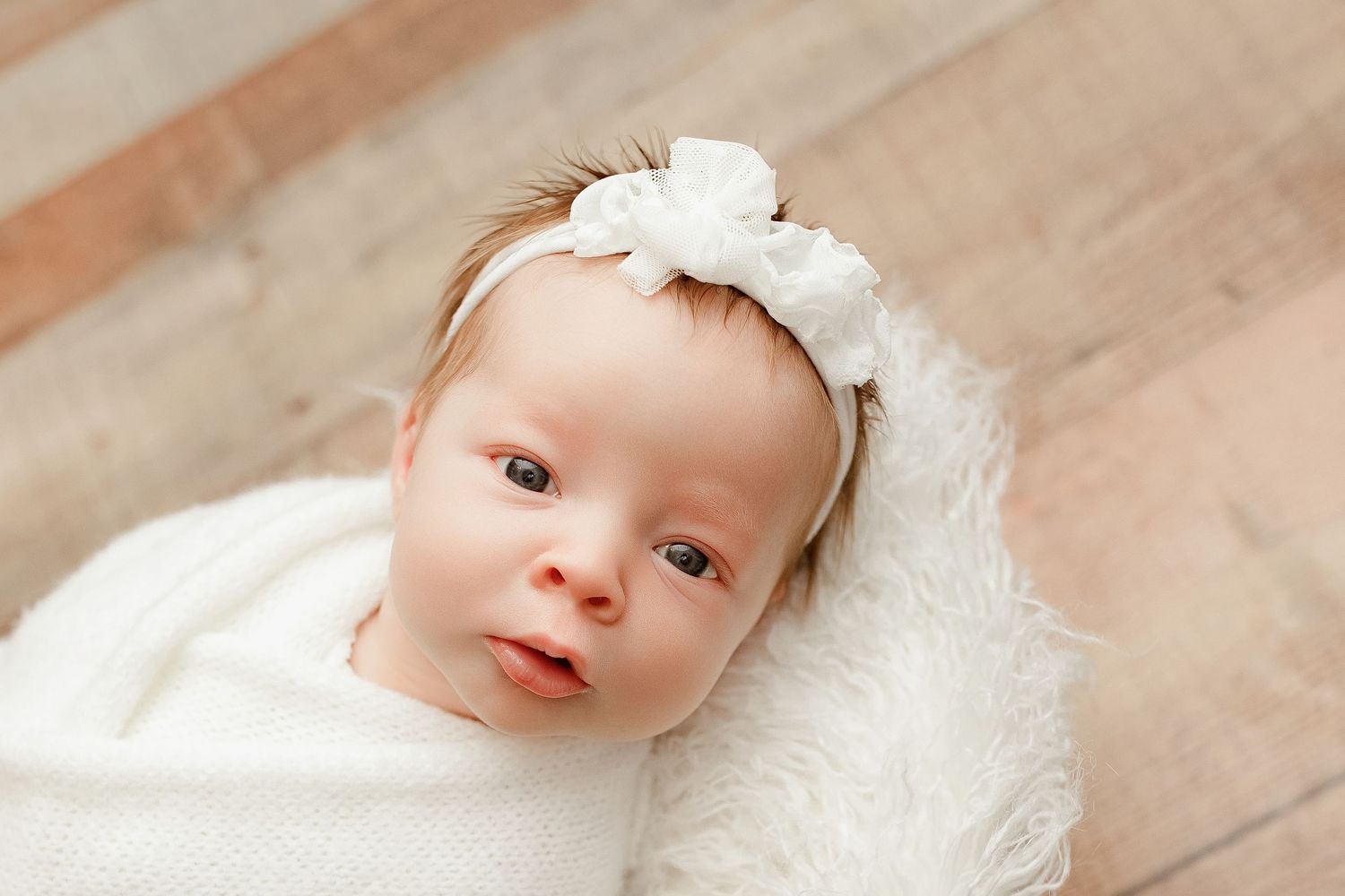 A baby in white clothing lies on a wooden floor while smiling and looking up at the camera.
