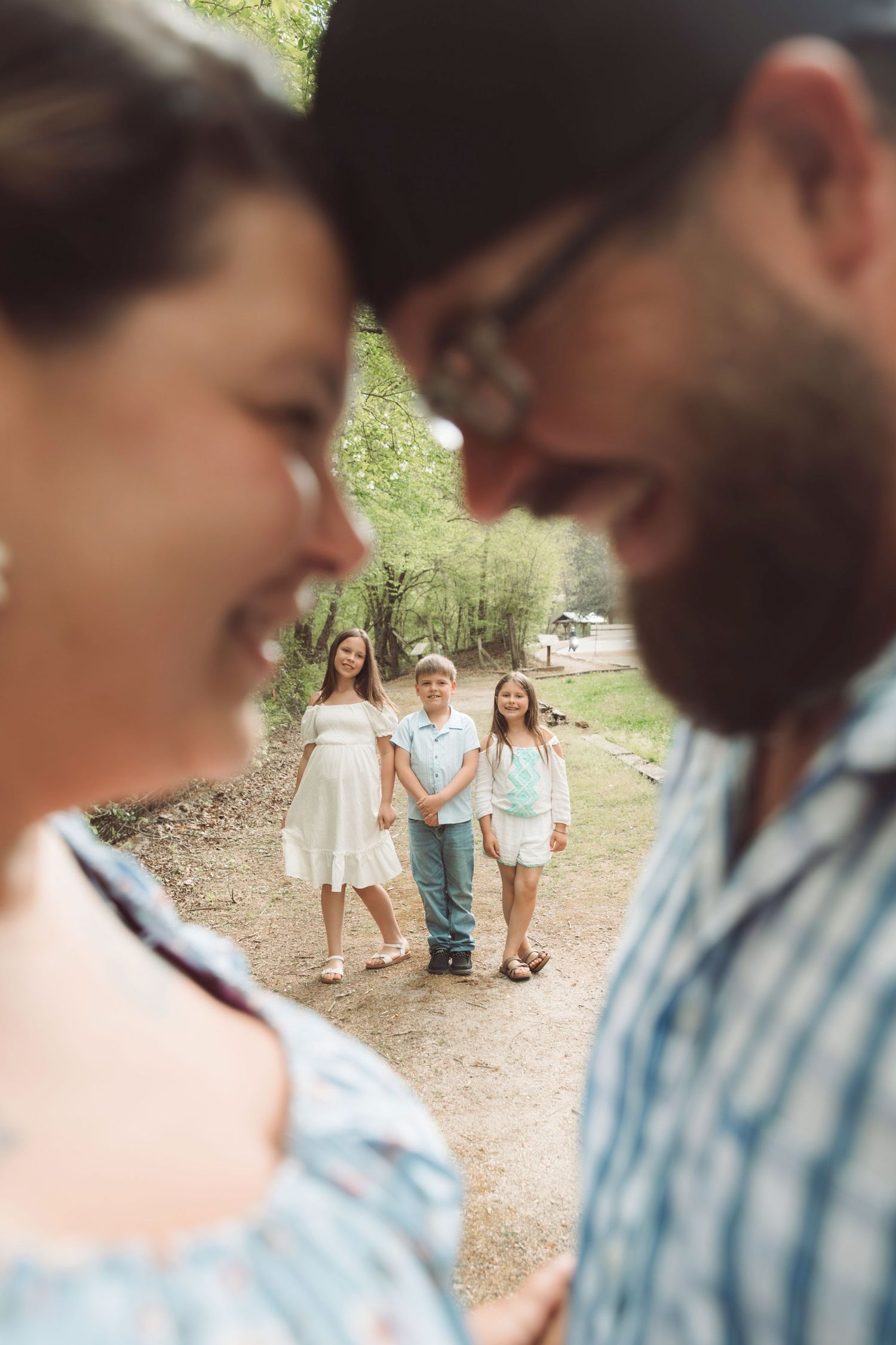 A couple laughs together in the foreground while three children smile and hold hands on a nature path behind them.