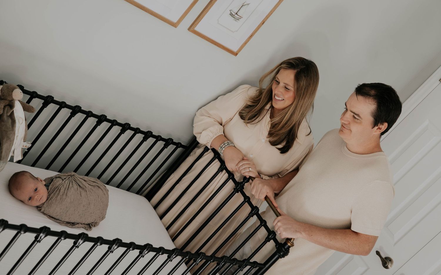 Two people stand by a wooden crib looking down at a swaddled baby in a neutral-toned nursery setting.