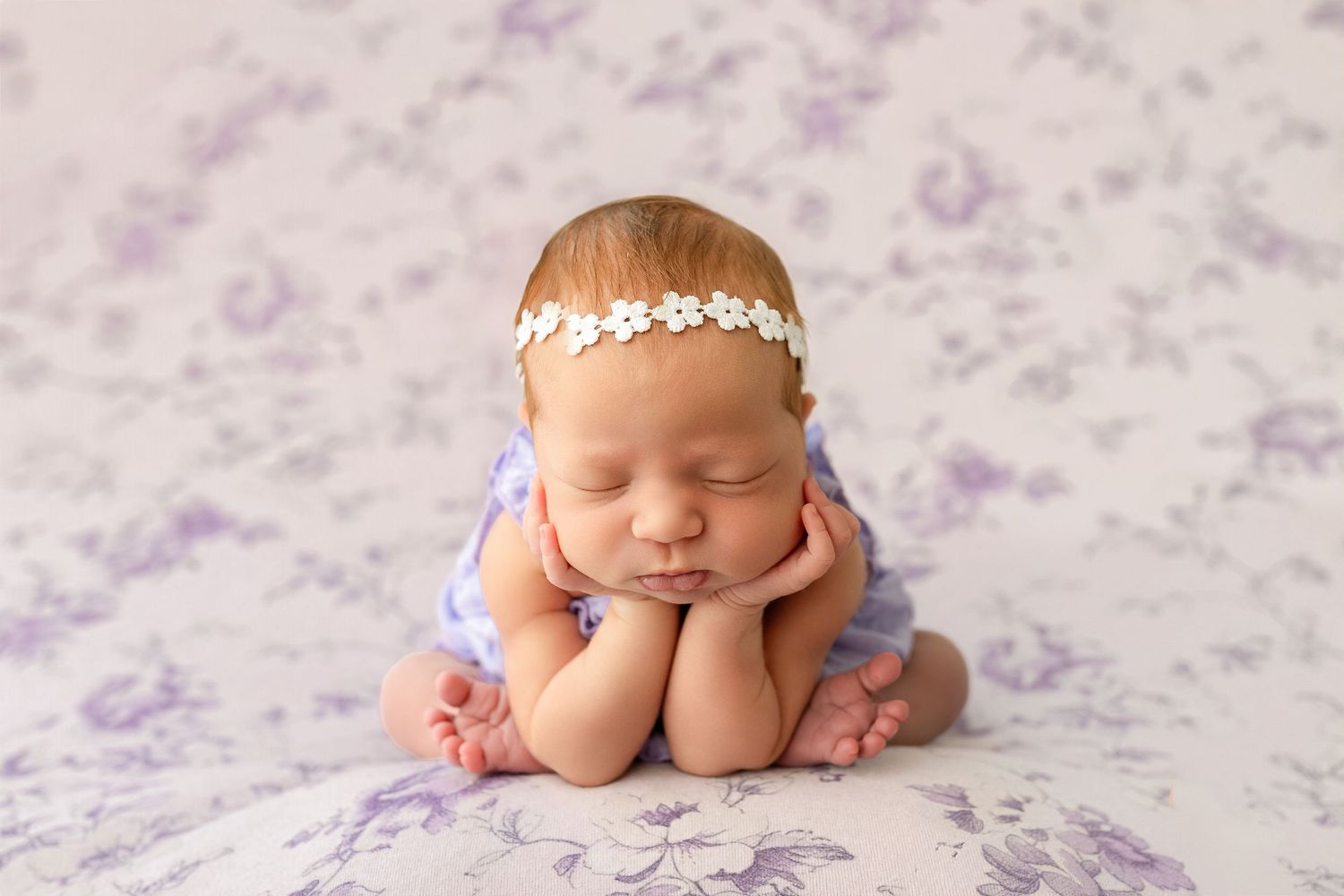 Sleeping newborn baby girl wearing a white flower headband posed on a purple floral blanket.