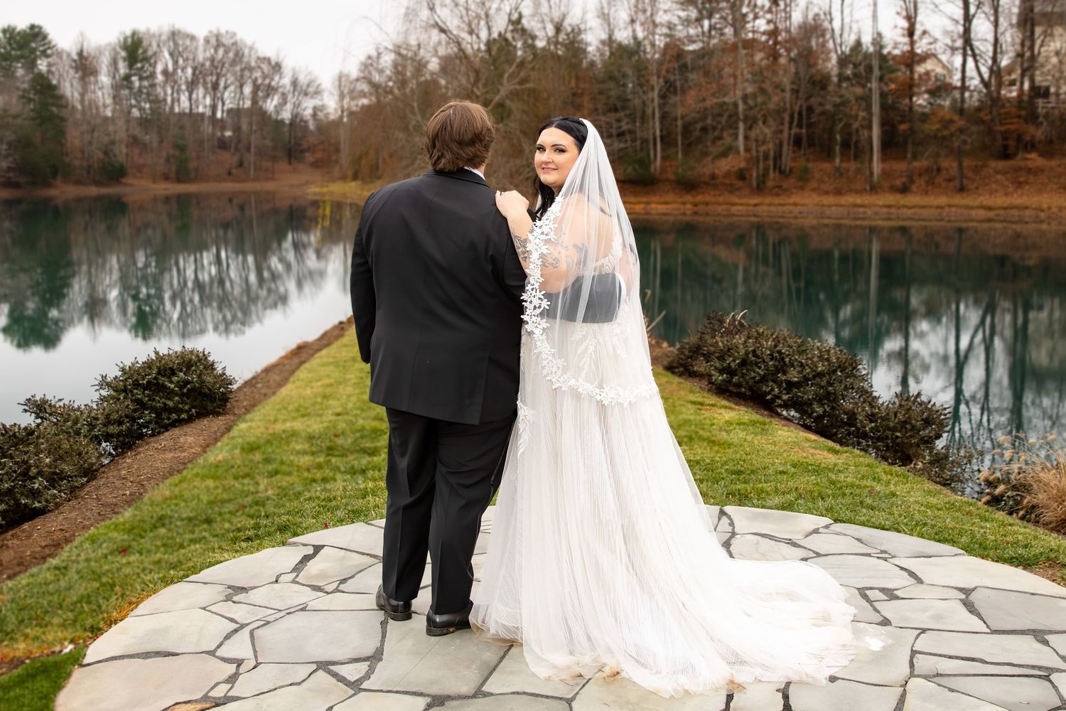Bride in lace gown and veil stands with groom in black tuxedo on stone patio beside serene lake.