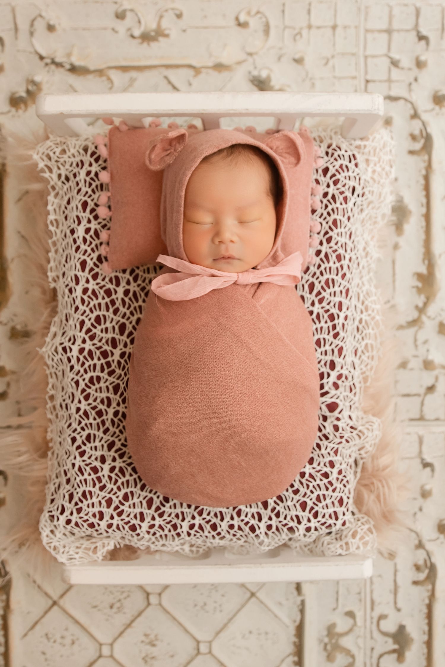 Newborn baby sleeping peacefully on delicate white lace pillow in soft peach tones during professional photo session.