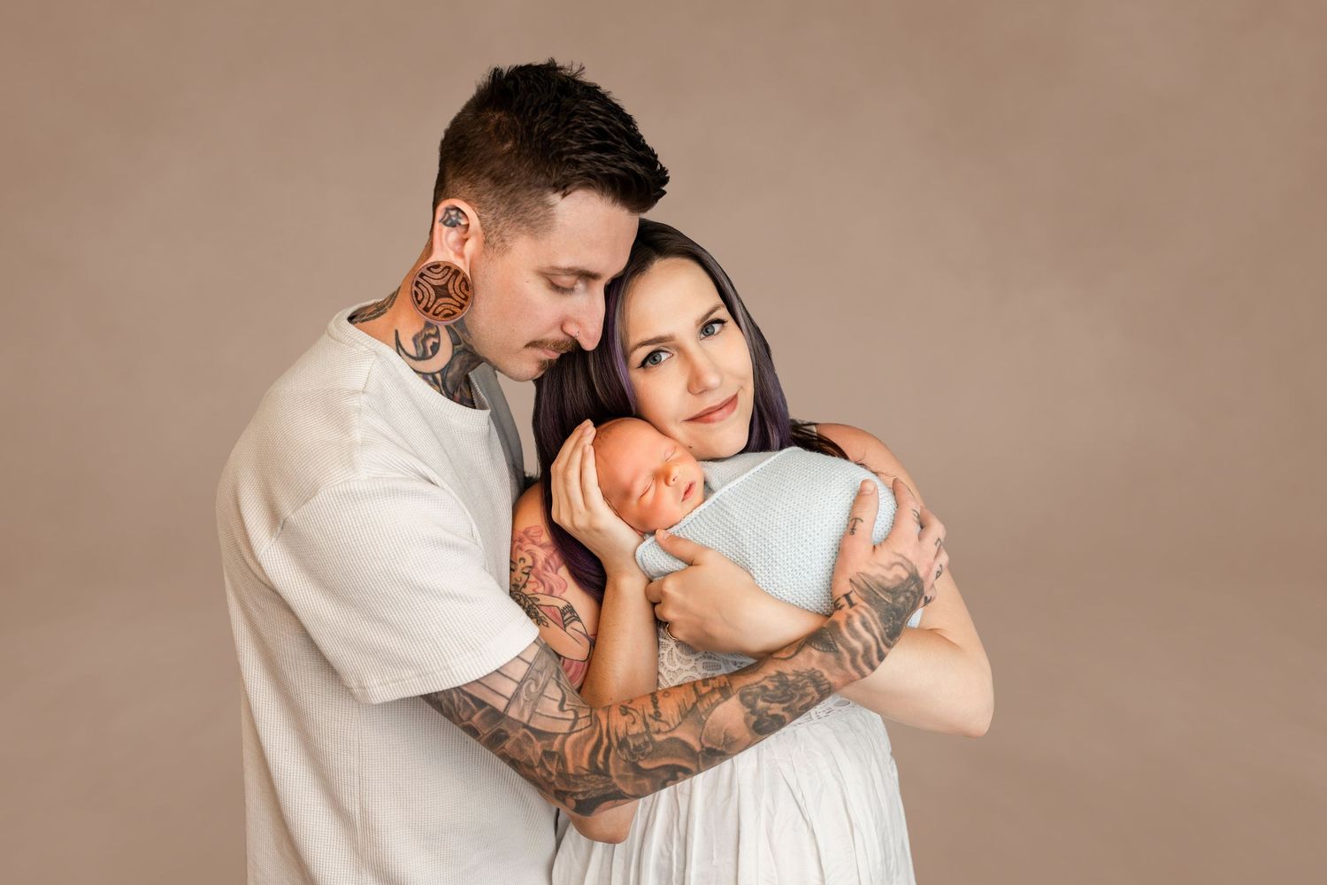 Tattooed parents tenderly hold their newborn baby during a studio portrait session against a warm beige background.